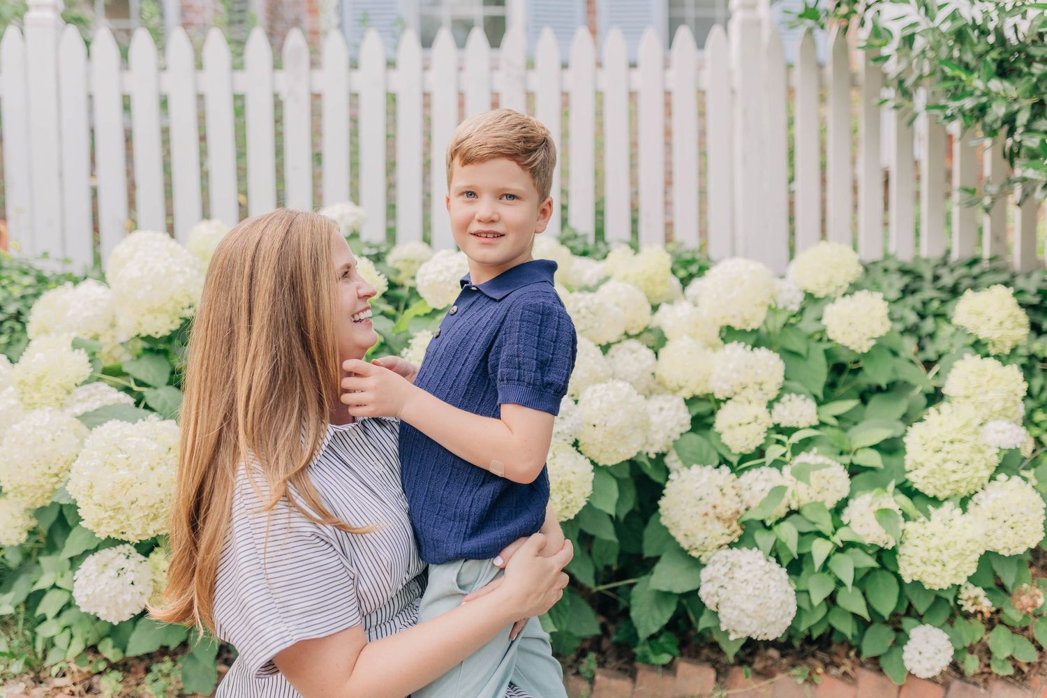 A woman lovingly holds a young boy in a navy polo near blooming white hydrangeas outdoors.