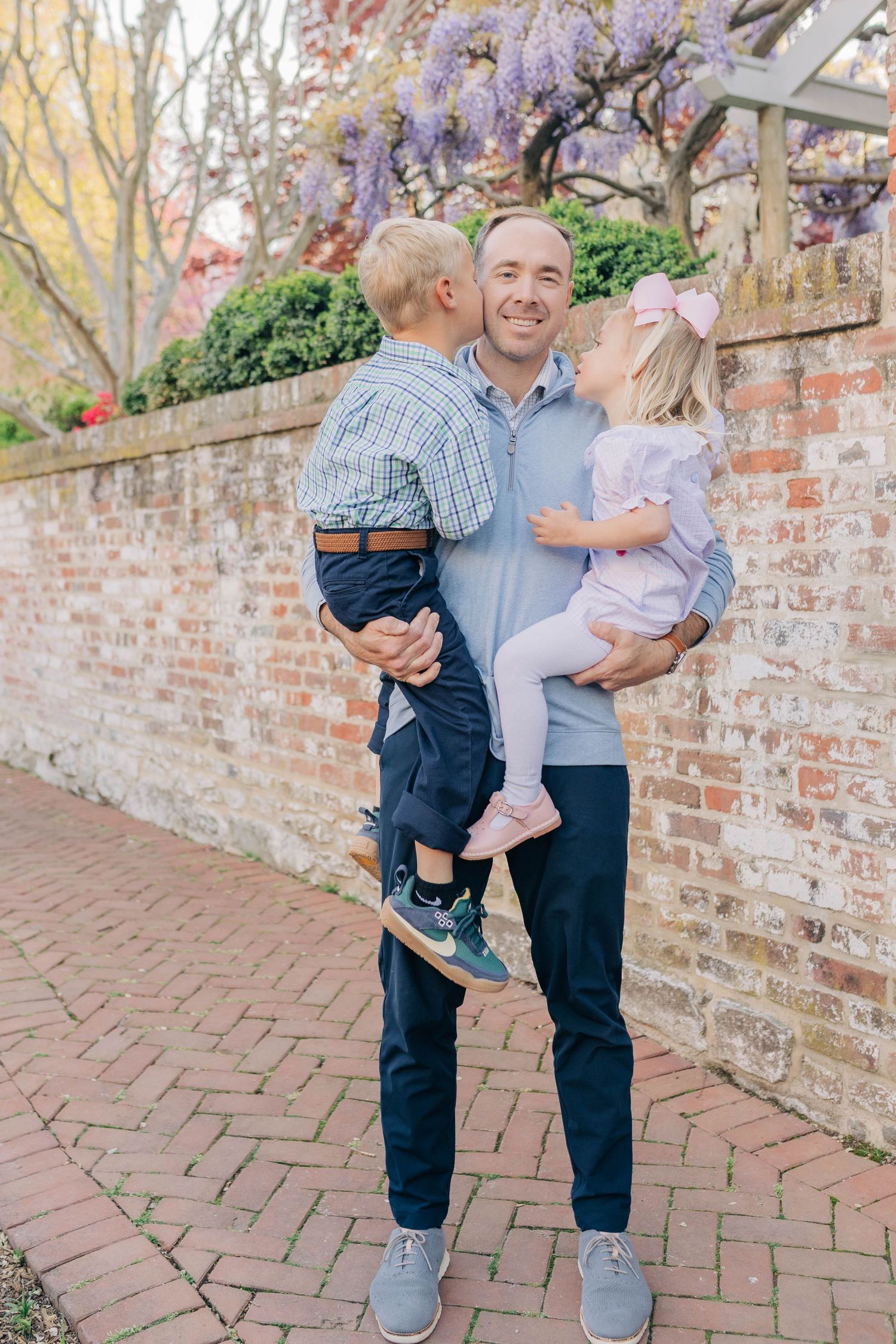 A smiling man holds two young children in front of a brick wall with blooming wisteria in a garden setting.