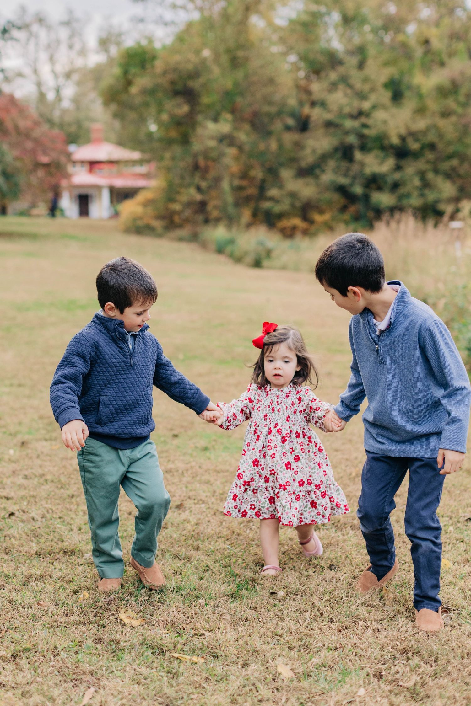 Two boys in blue sweaters hold hands with a little girl in a floral dress outdoors in a green park.