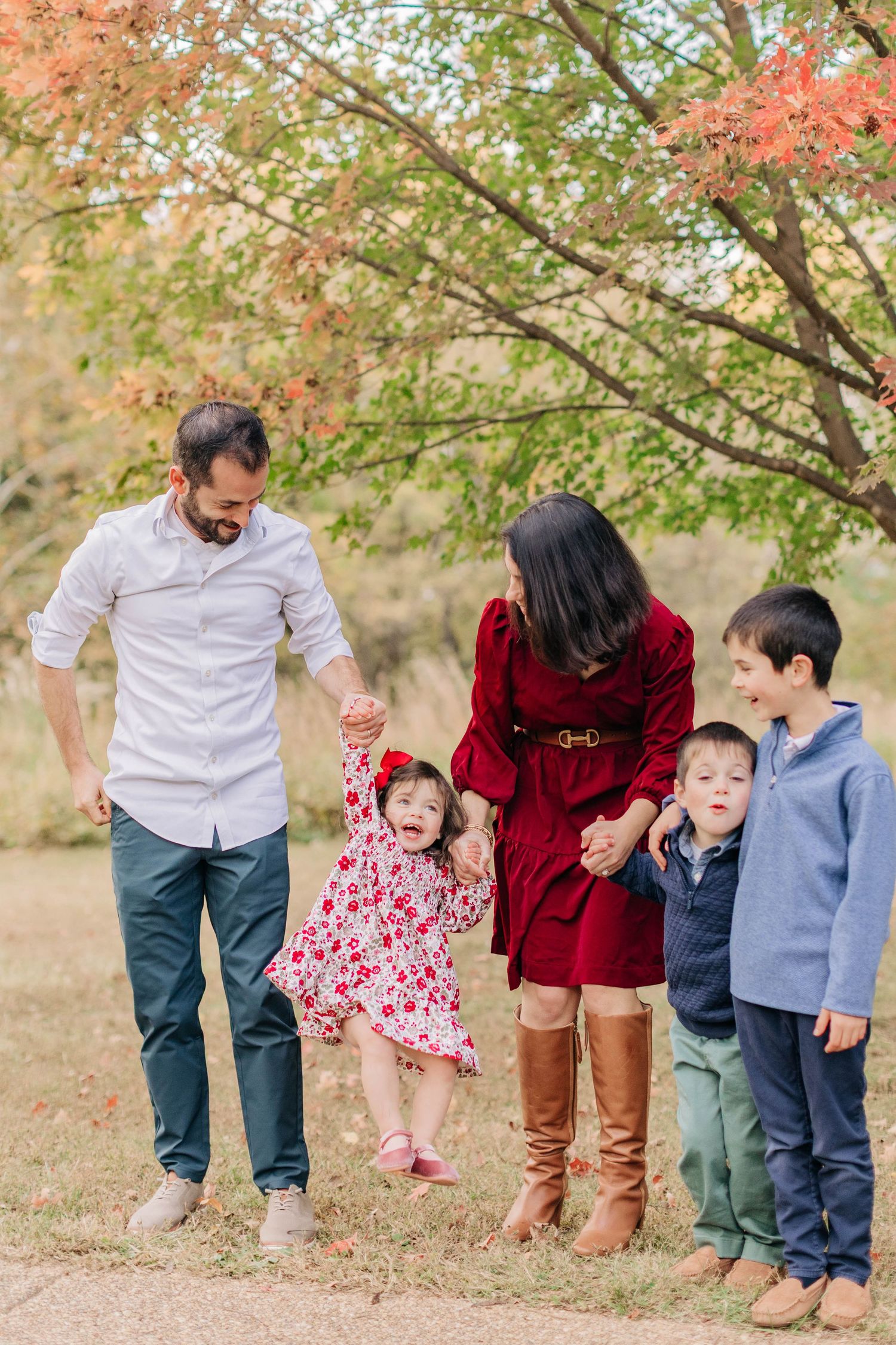 Family of five enjoying autumn park outing, parents swinging toddler girl while two boys watch, fall foliage background.