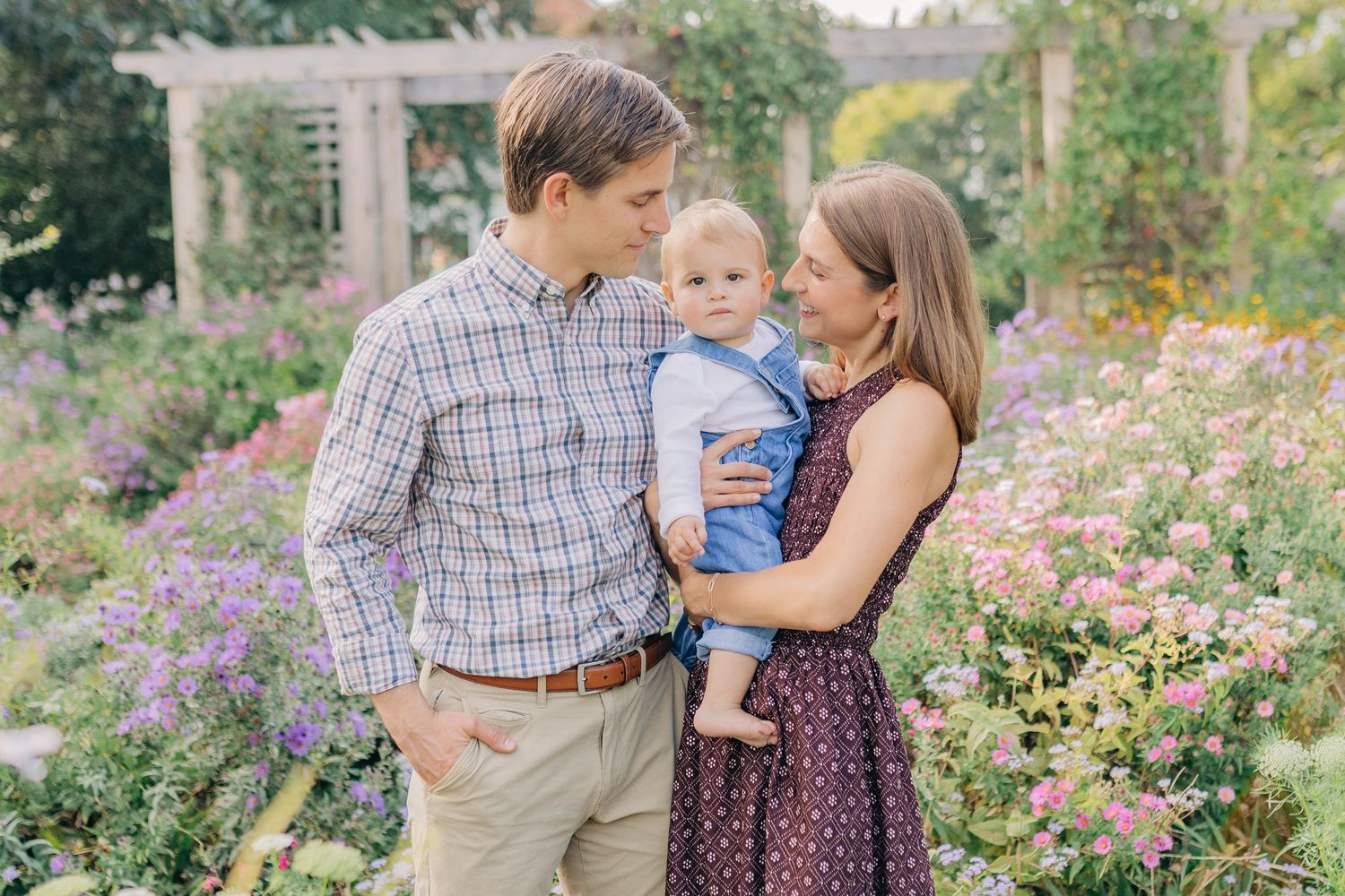 A young family with a baby boy poses among colorful blooming flowers in a lush garden setting.