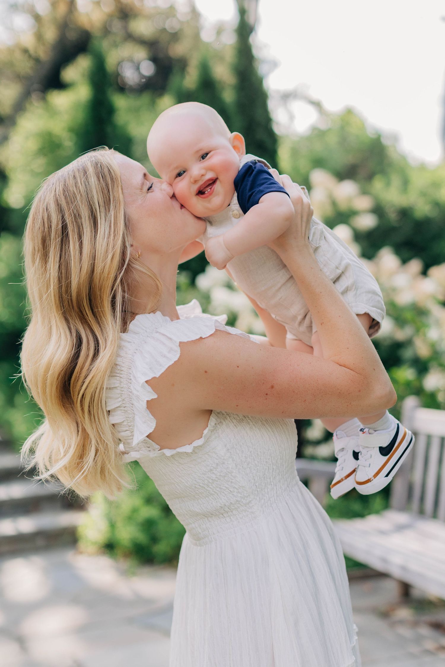 Loving mother in white dress kissing and lifting her laughing baby outdoors.