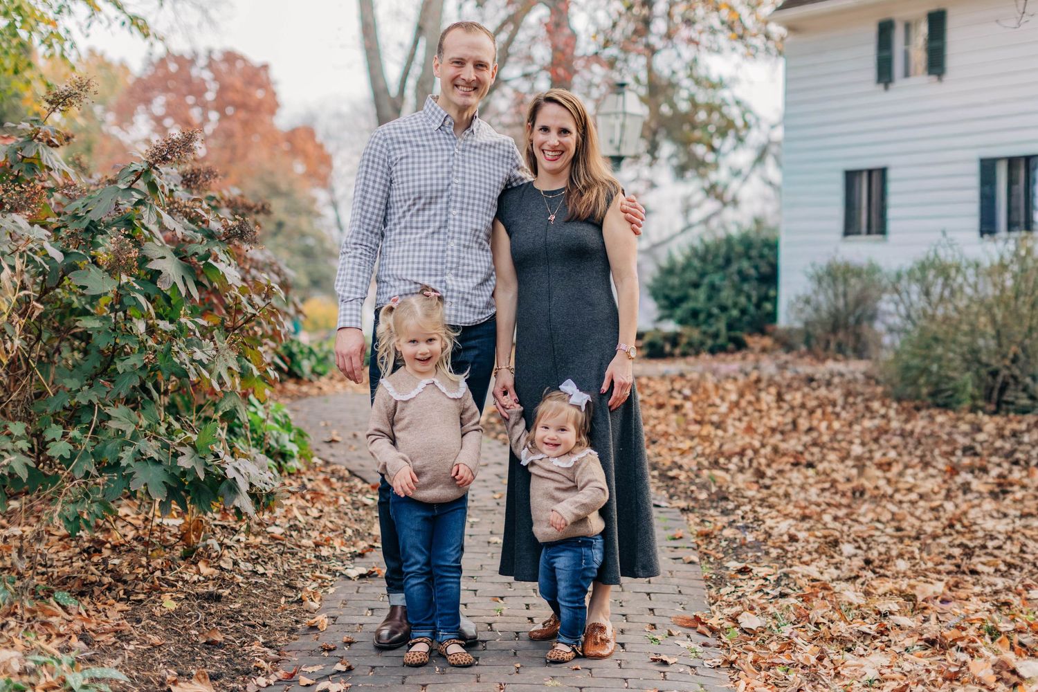 Family of four posing outdoors in autumn, parents with two toddlers on a leaf-covered path near a white house.