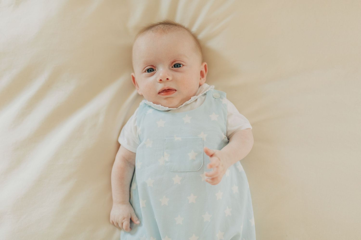 Baby wearing light blue outfit lies on white bedding while looking at camera.