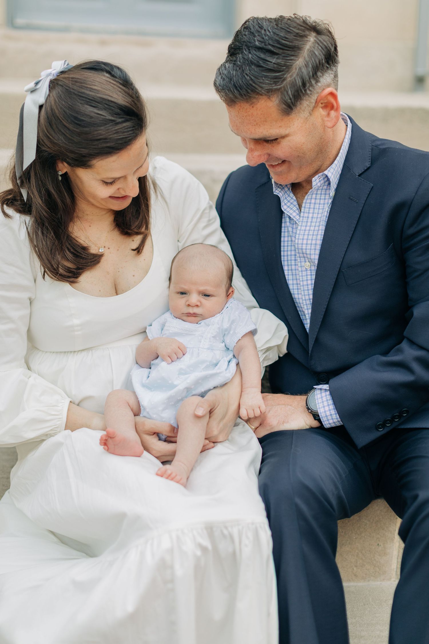 Parents in formal wear sit with baby on stone steps for portrait.
