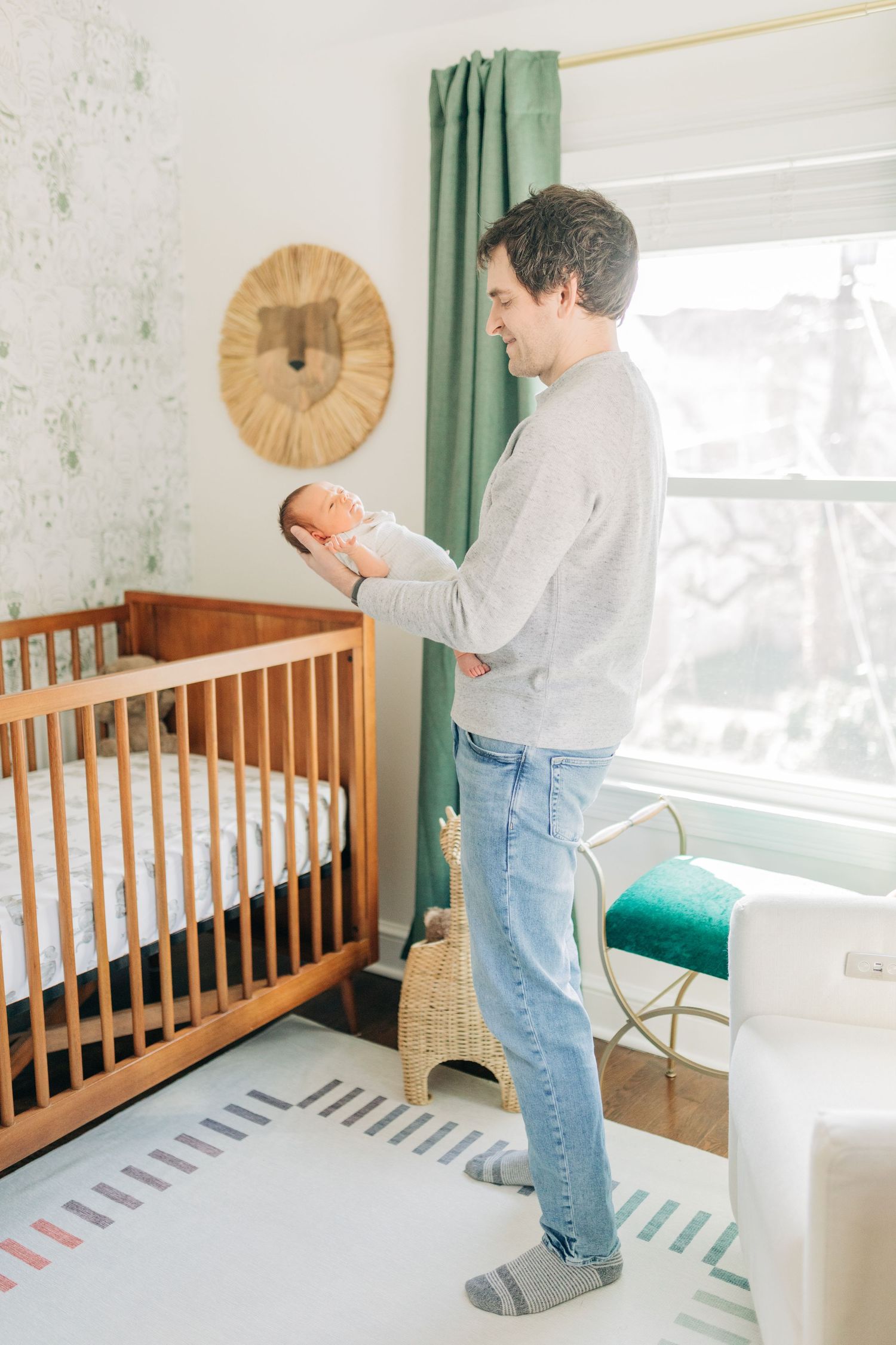 Parent holding infant while standing near wooden crib in sunlit nursery with green curtains.