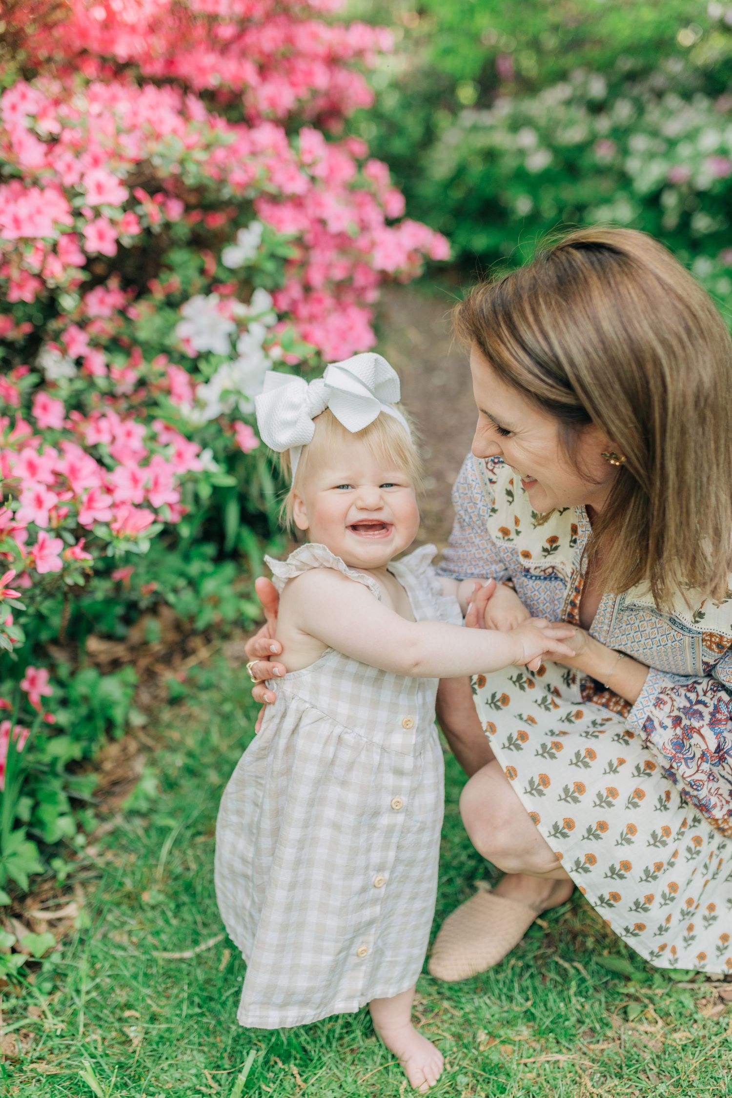 Mother and baby sharing joyful moment among pink flowering bushes in garden setting.