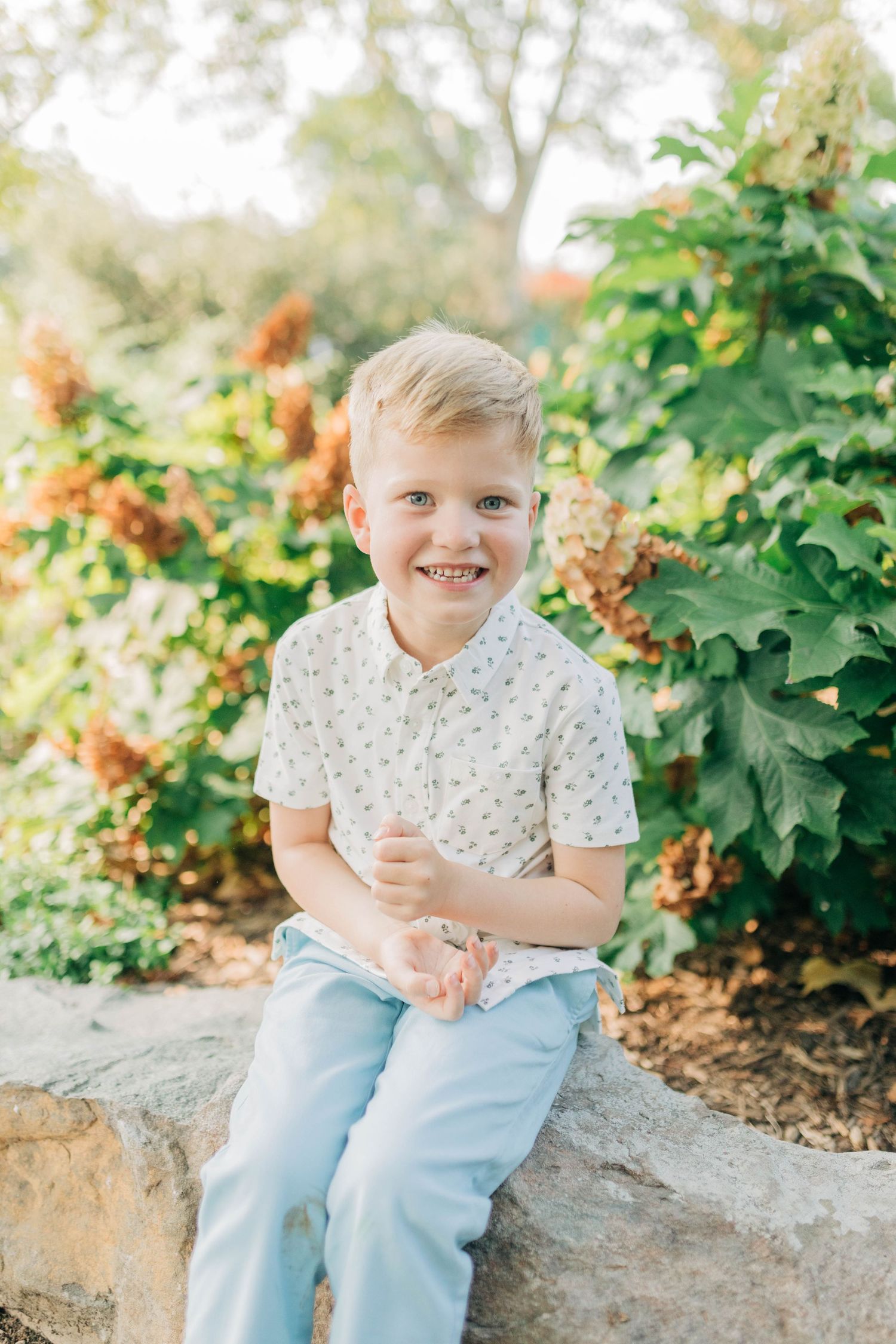 Child wearing light colored shirt and jeans sits casually on stone wall in garden setting.