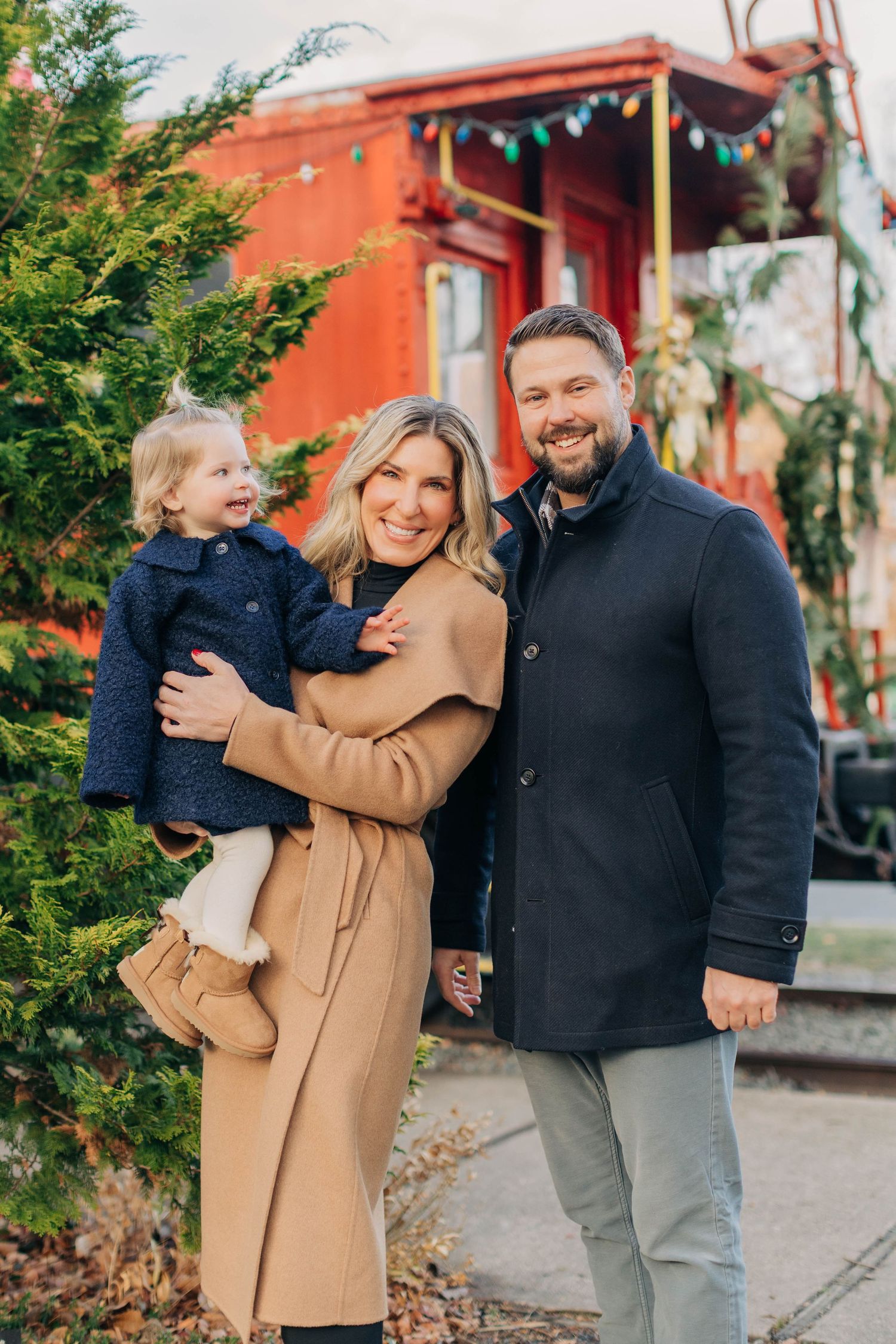 A smiling family of three poses outdoors in winter coats near a red train car and evergreen trees.