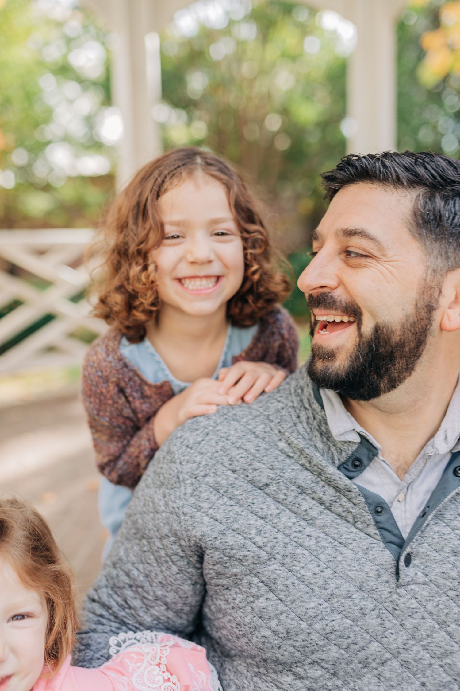 A bearded man laughs joyfully with two young curly-haired girls outdoors near a white pergola.
