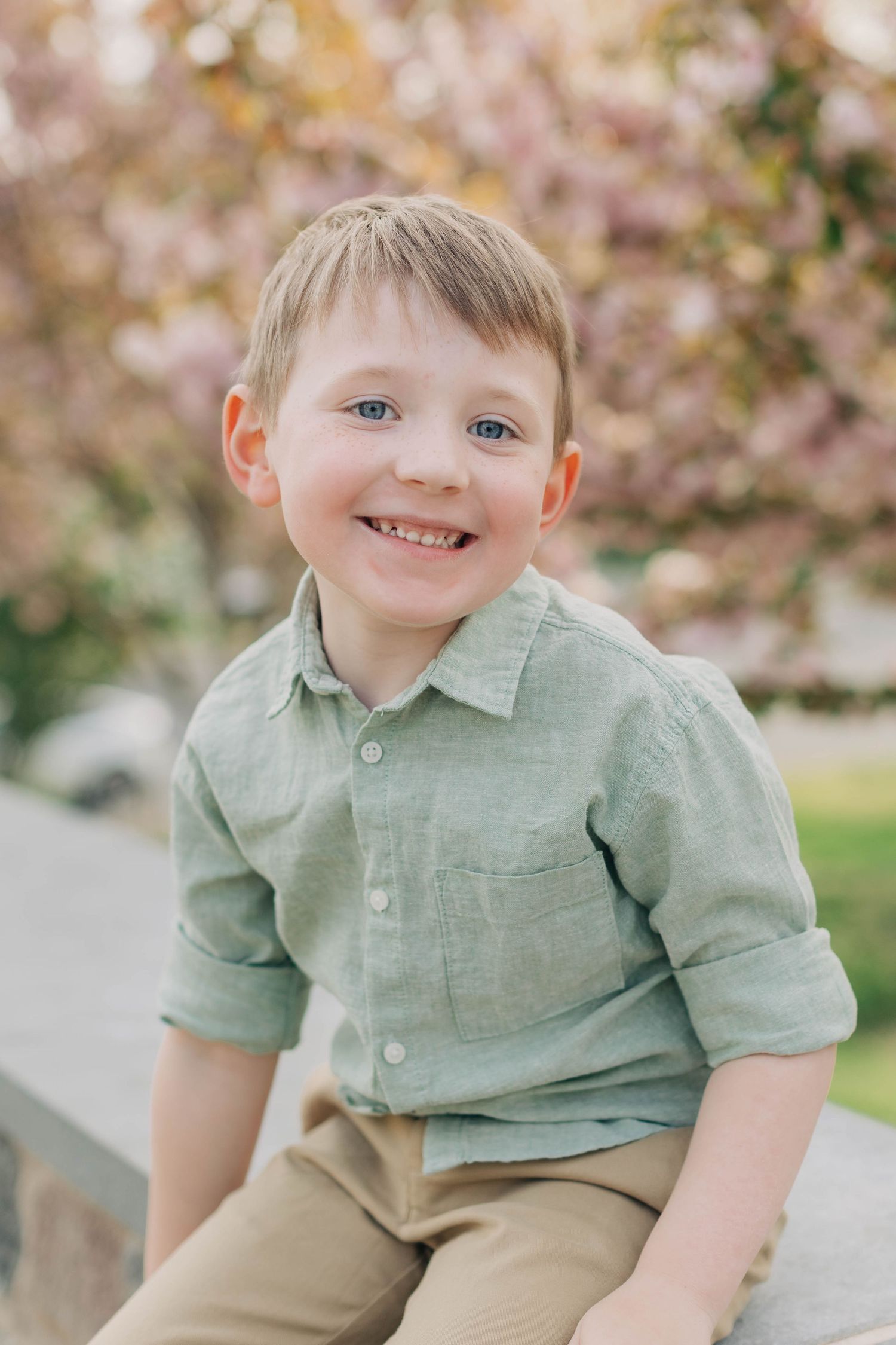 Smiling young boy with blue eyes wearing a green linen shirt sitting outdoors near pink cherry blossom trees.