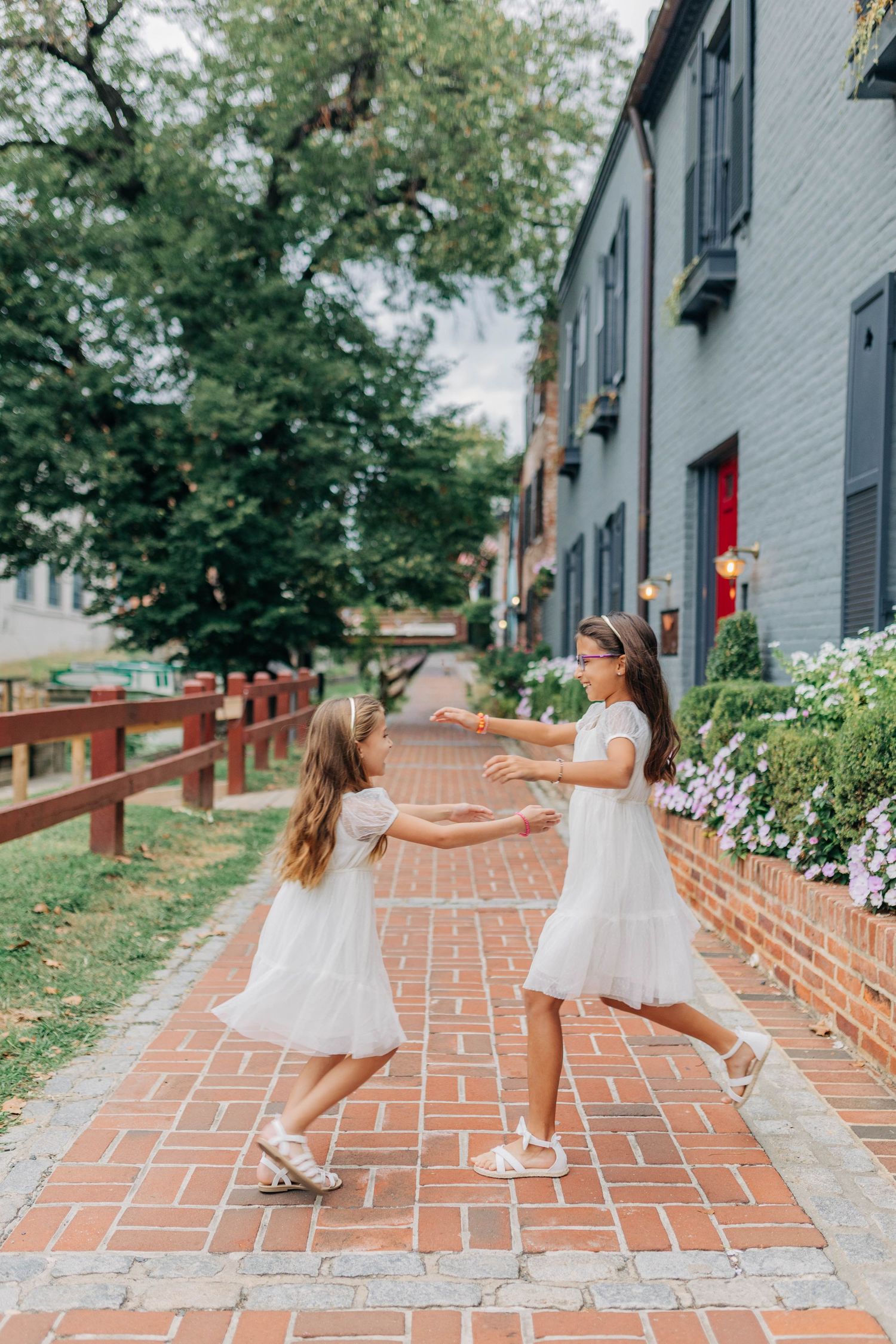 Two girls in white dresses playing on a brick pathway beside a gray building with flowers.