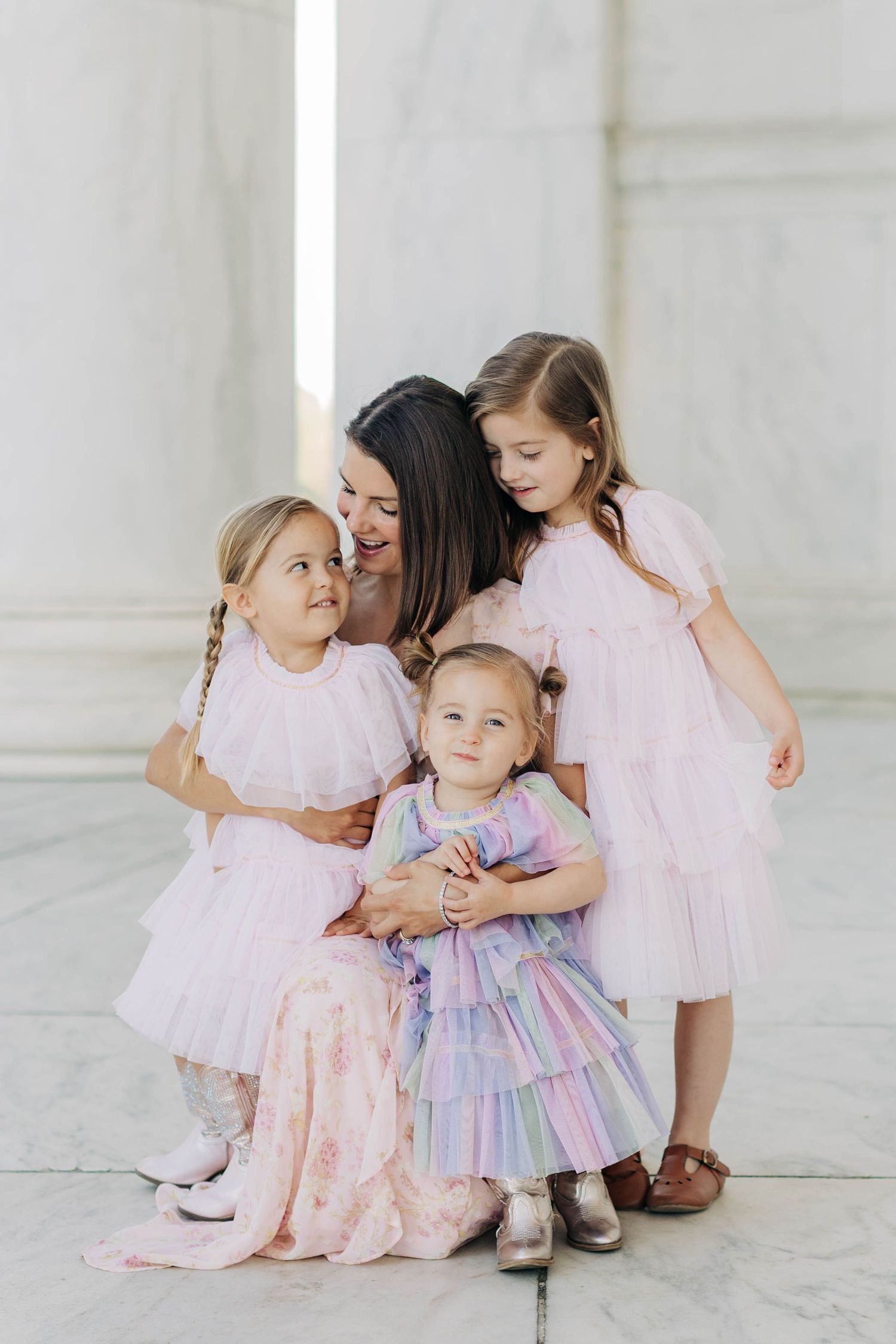 A mother in a pink floral dress poses with three young daughters in pink tutus at a marble monument.