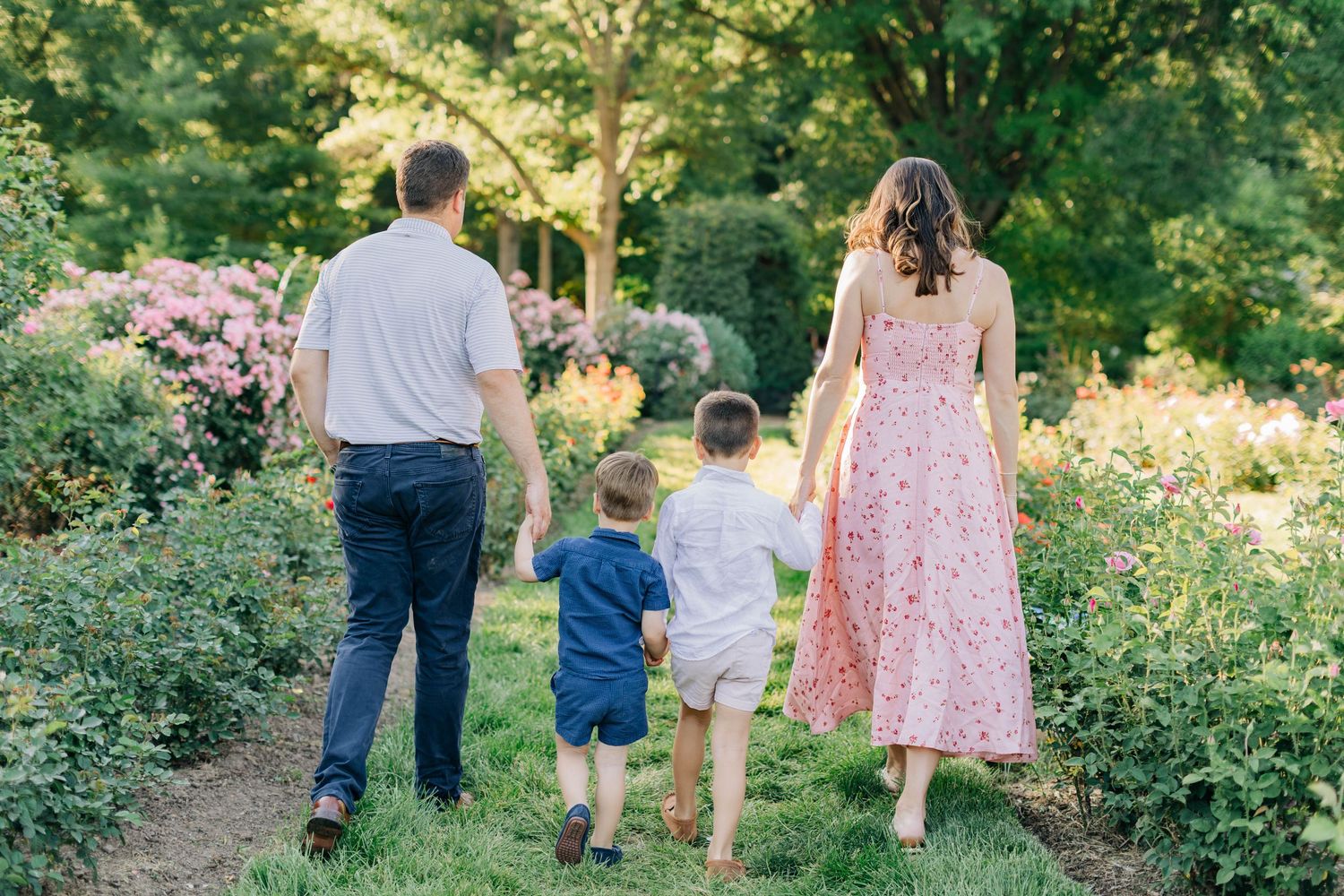A family of four walks hand-in-hand along a lush garden path surrounded by colorful blooming flowers.