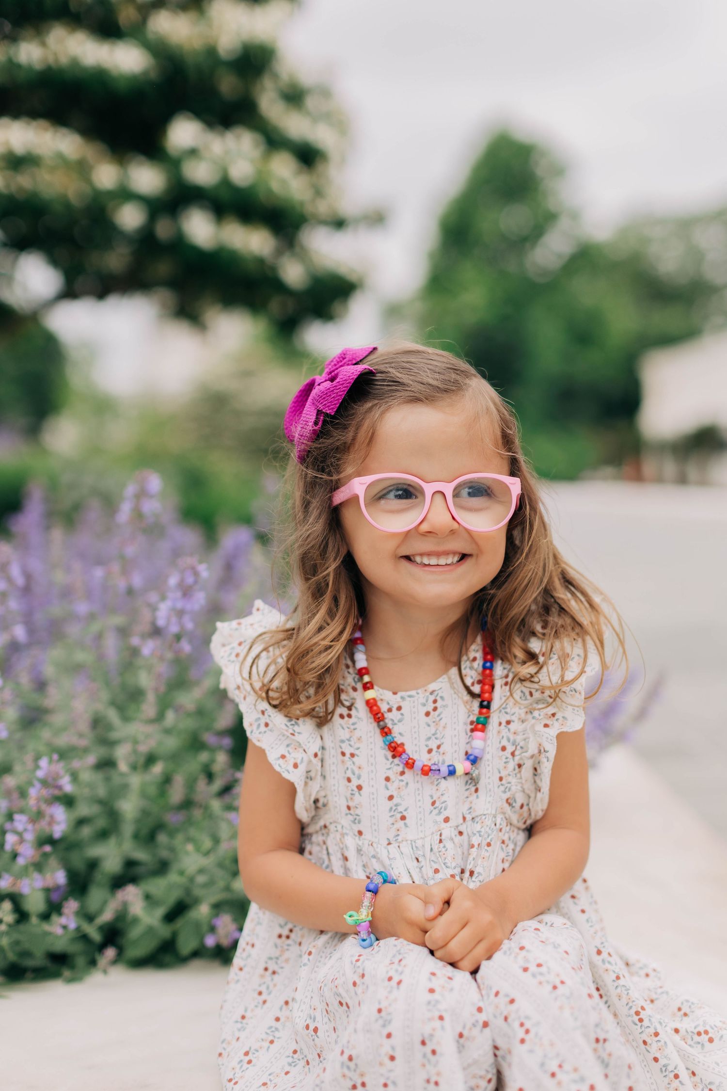 A smiling little girl wearing pink glasses, a floral dress, and colorful beaded necklace poses outdoors near purple flowers.