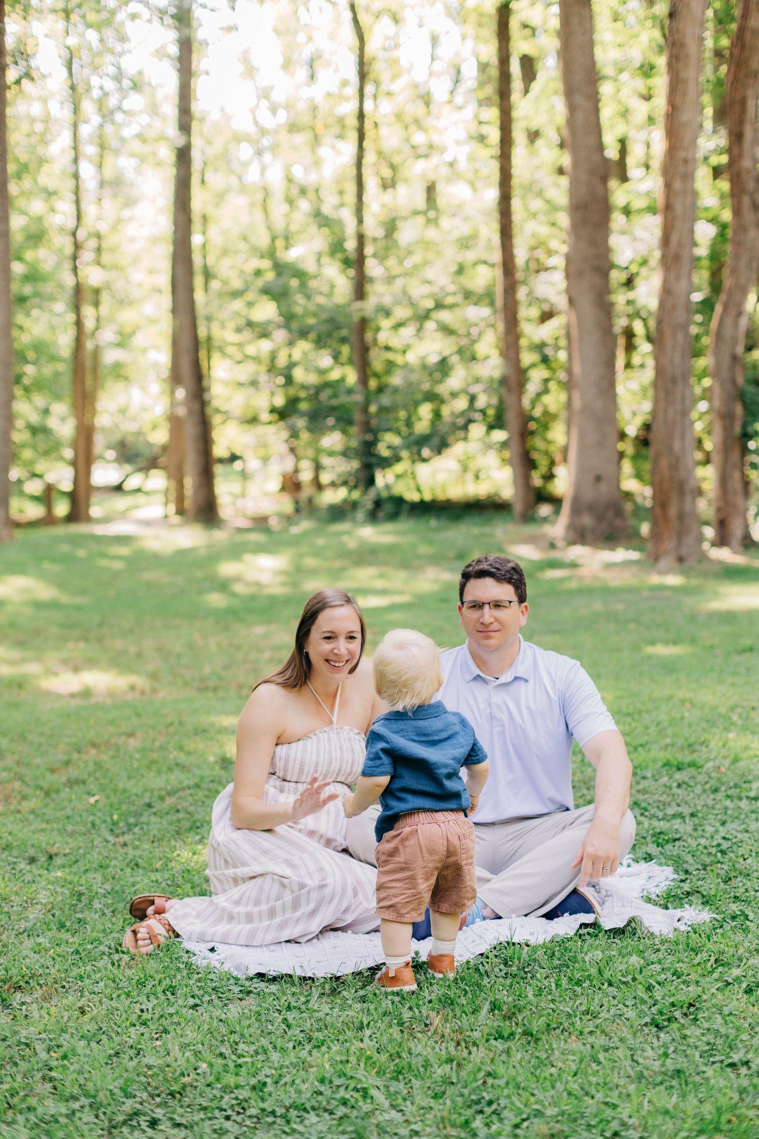 Family of three sitting on a blanket in a sunny park, parents smiling at toddler standing between them.