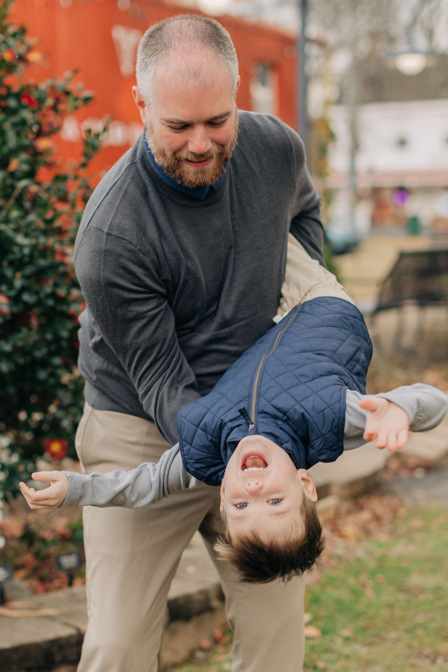 A bearded father playfully holds his laughing child upside down outdoors, both smiling joyfully.