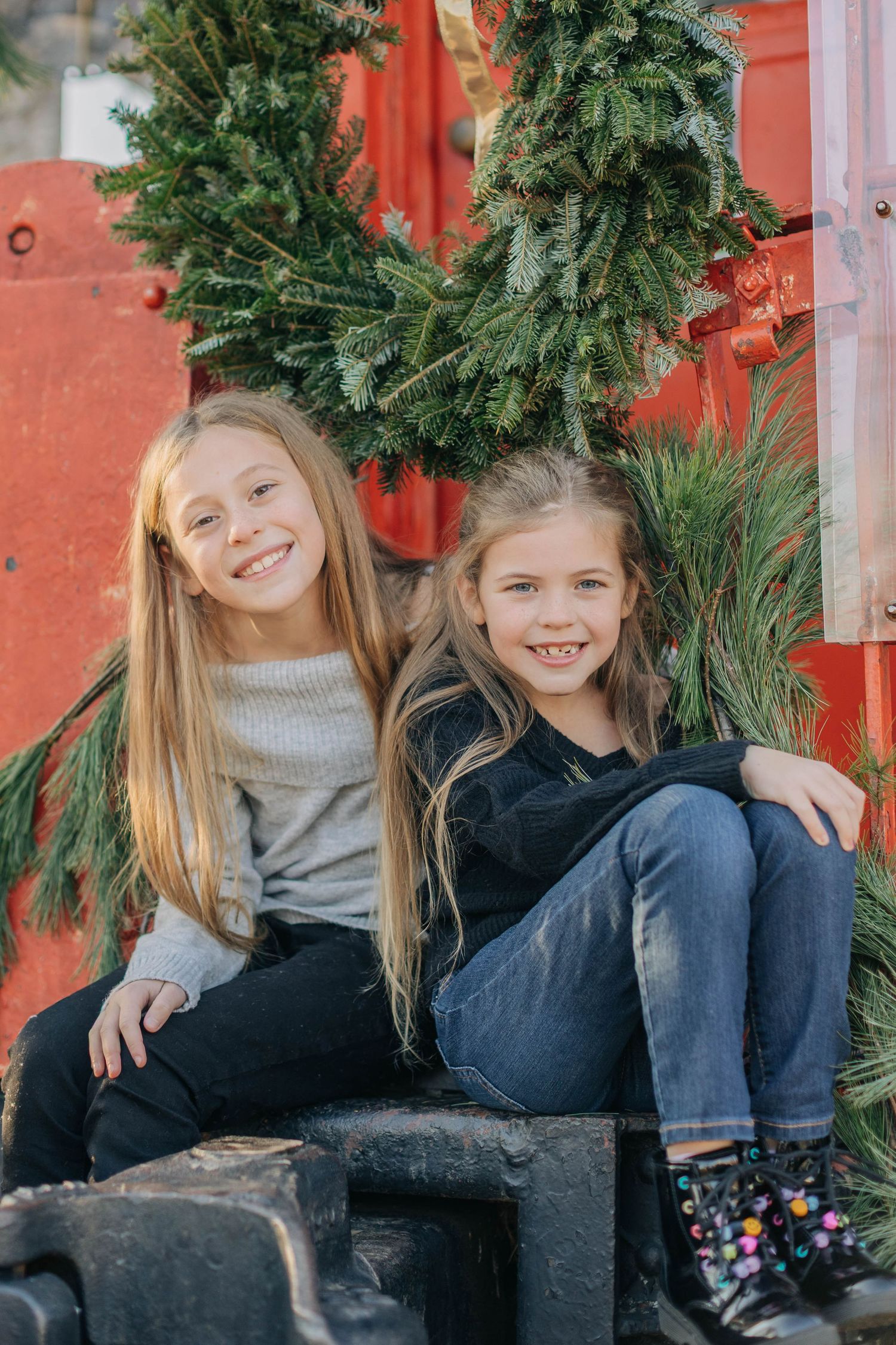 Two smiling girls with long hair sit together outdoors near a festive Christmas wreath and red barn backdrop.