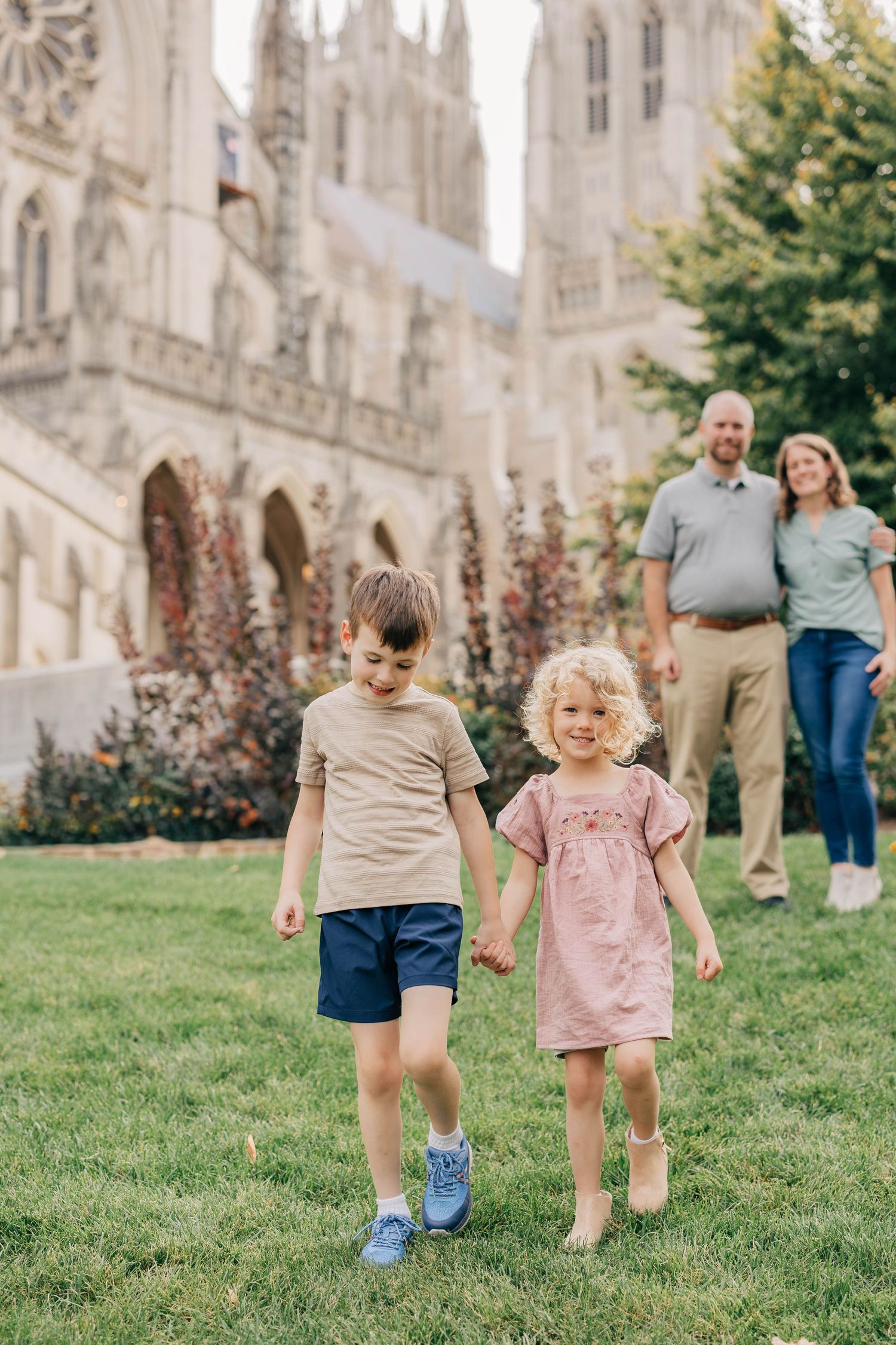Two young children hold hands and walk across a grassy lawn while their smiling parents stand nearby in front of a Gothic stone cathedral.