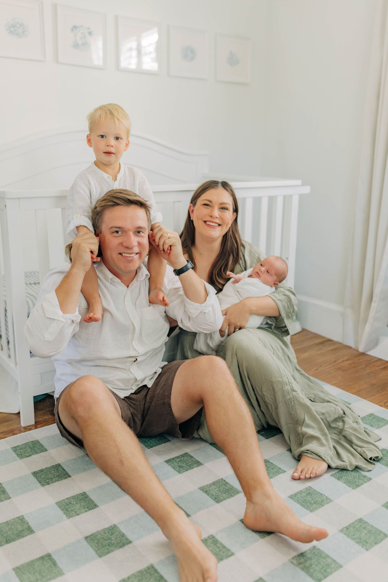 A family sits together on a checkered rug in a nursery with framed artwork on the wall.