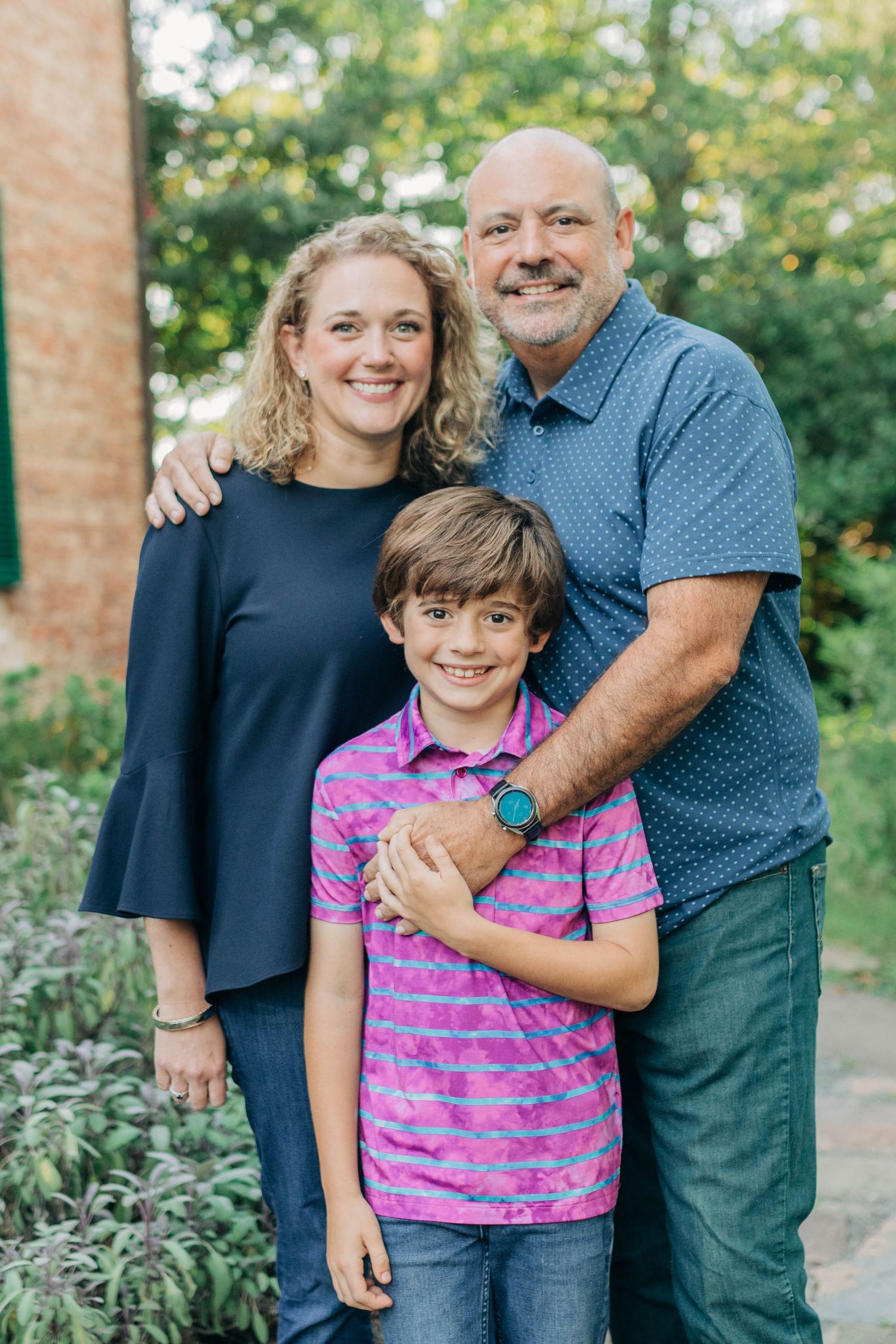 Three people share a warm embrace while standing outdoors near a brick building with lush landscaping.