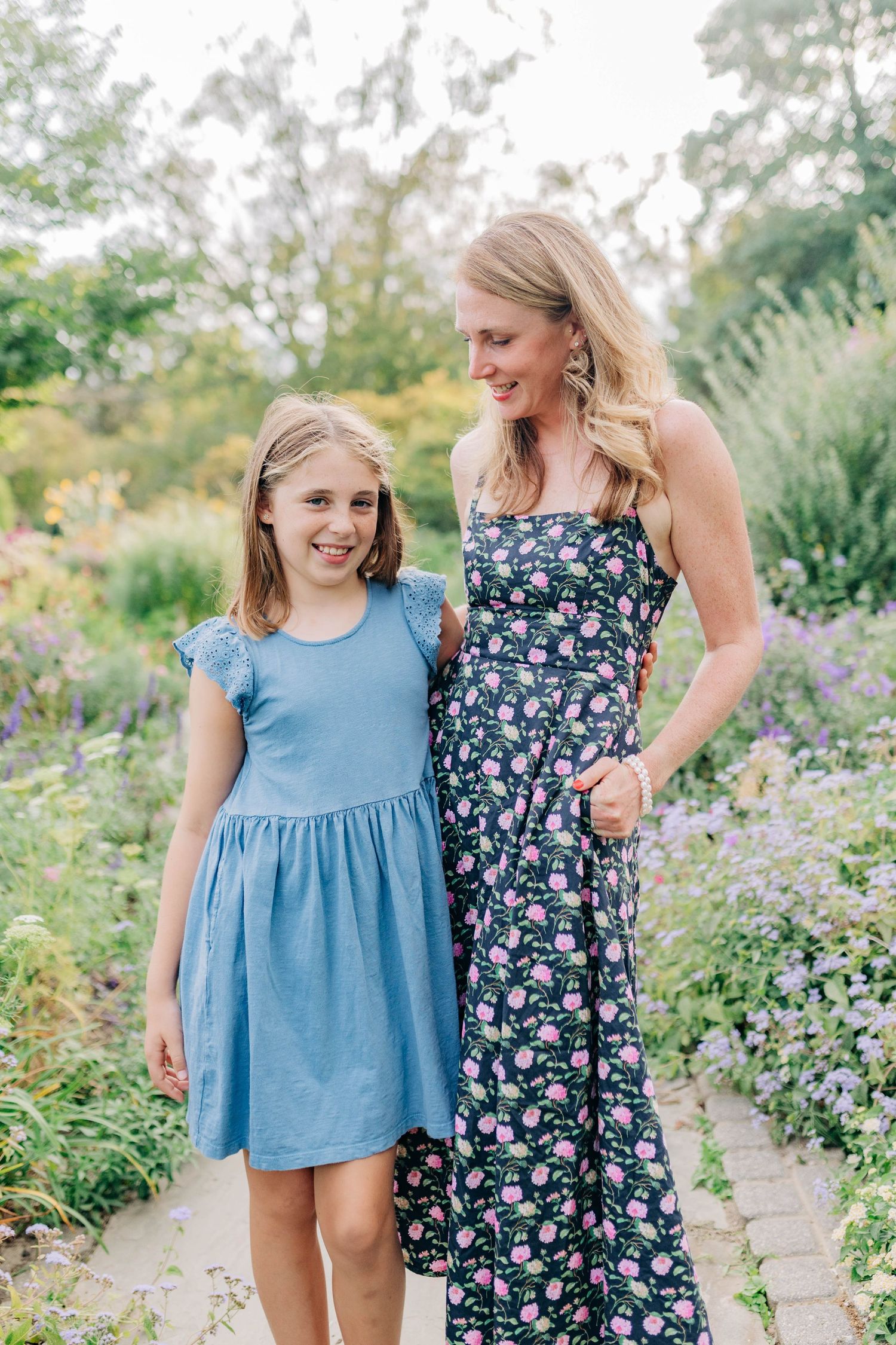 Two people in contrasting dresses stand together in a vibrant garden setting with purple flowers.