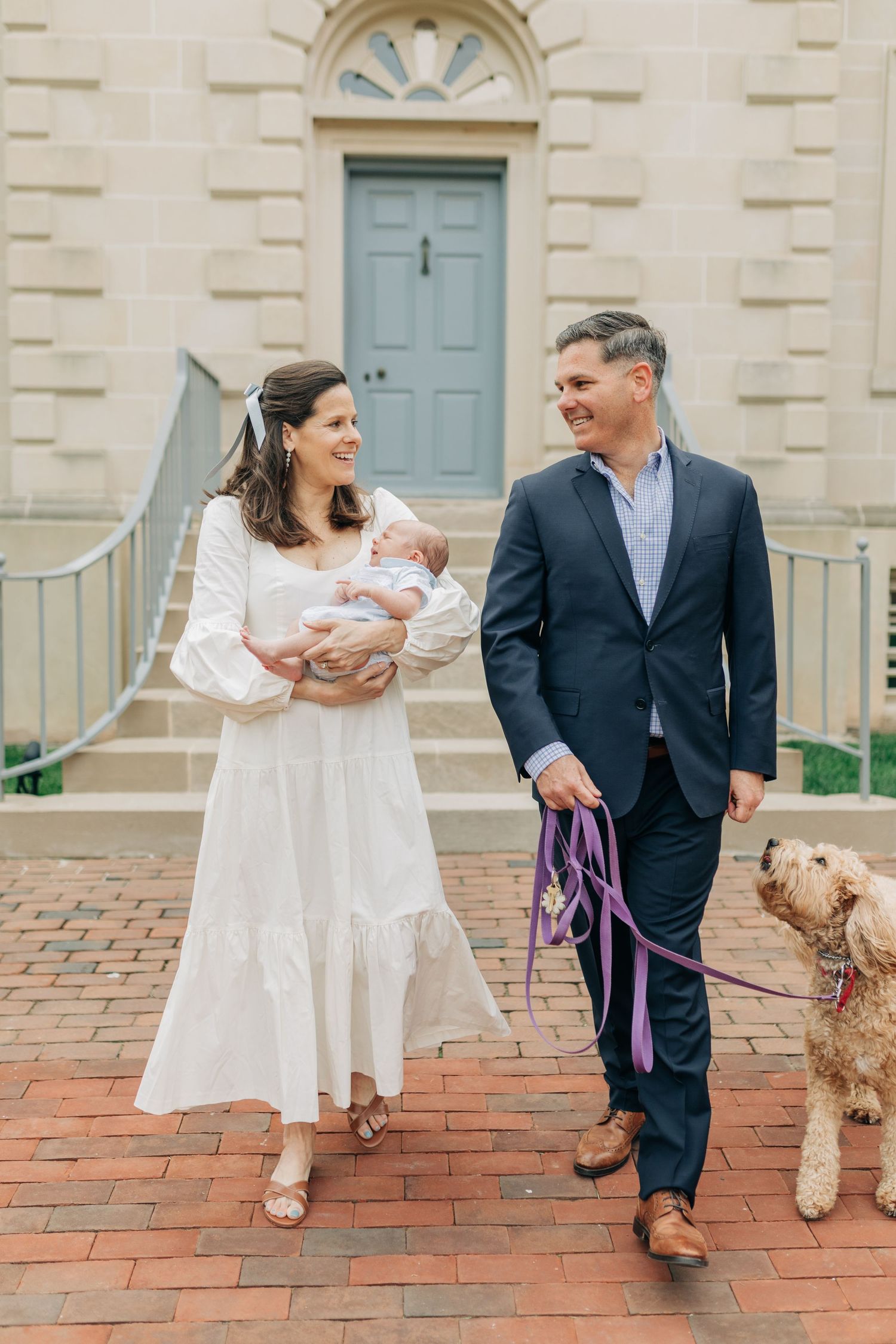 Family walking with their goldendoodle dog on brick patio near stone building.
