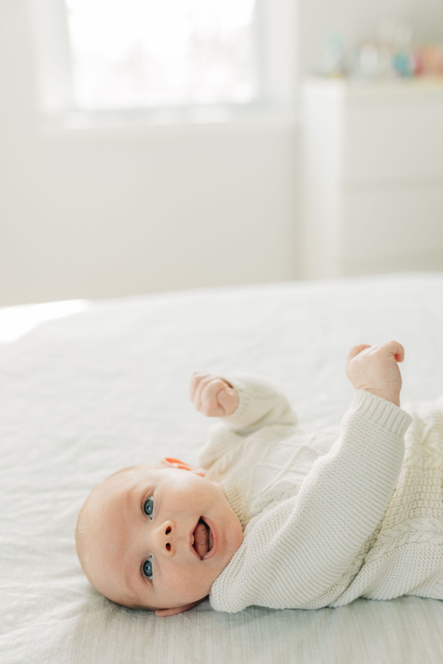 Baby wearing white cable knit sweater lying on white bed linens.