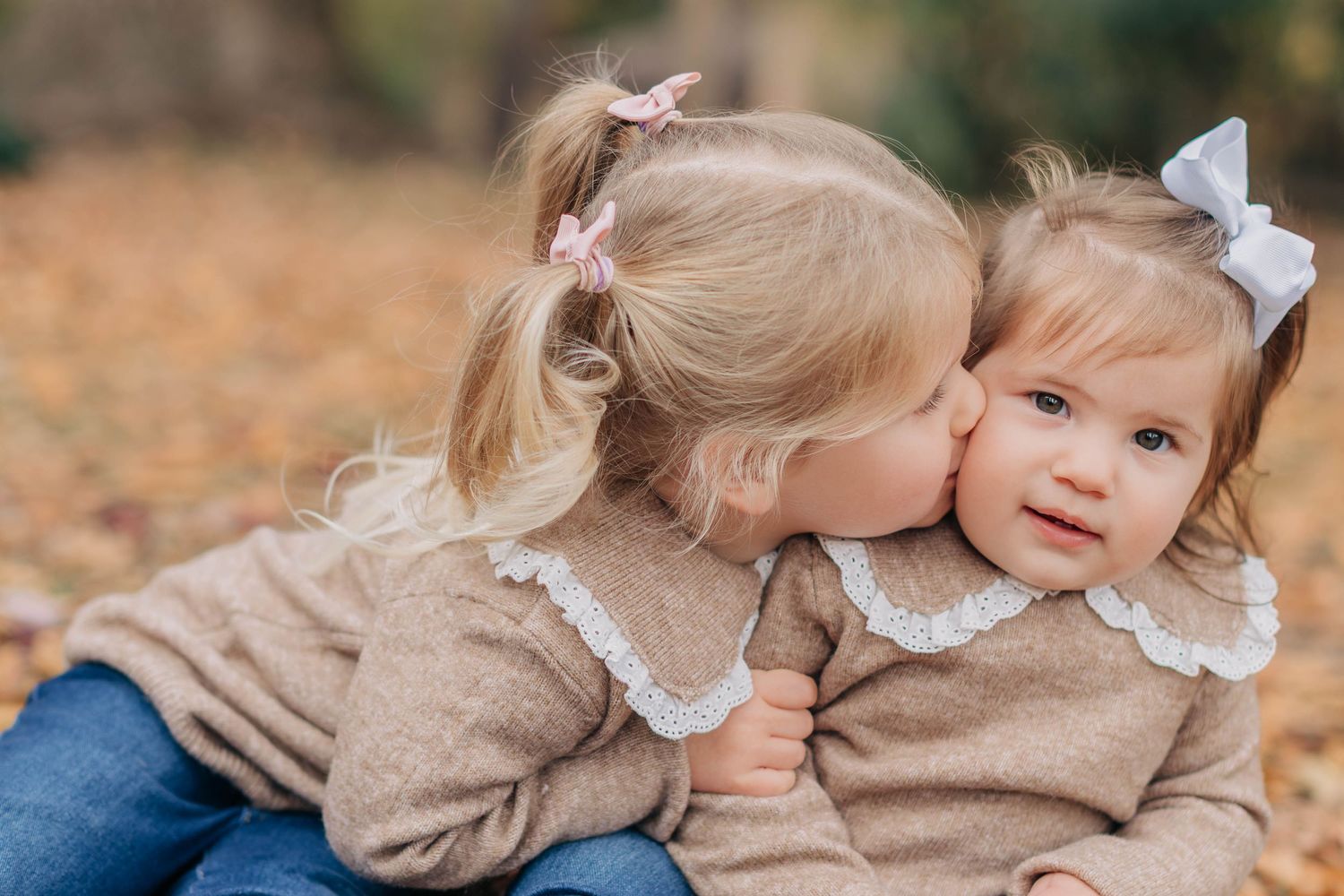 Two young sisters in matching beige sweaters share a sweet kiss outdoors among autumn leaves.