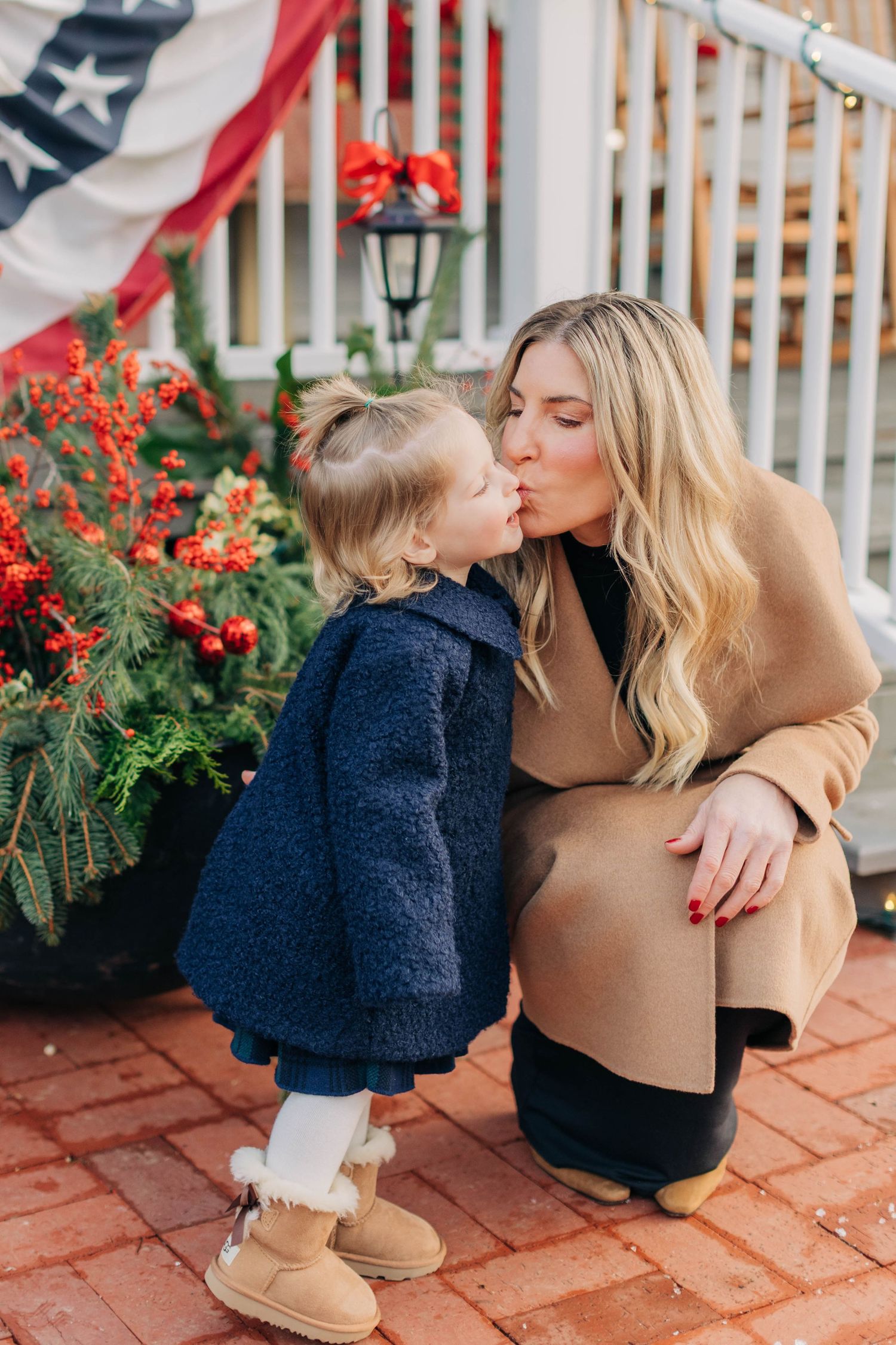 A blonde woman in a tan coat shares a sweet kiss with a toddler in a navy coat near holiday decorations.