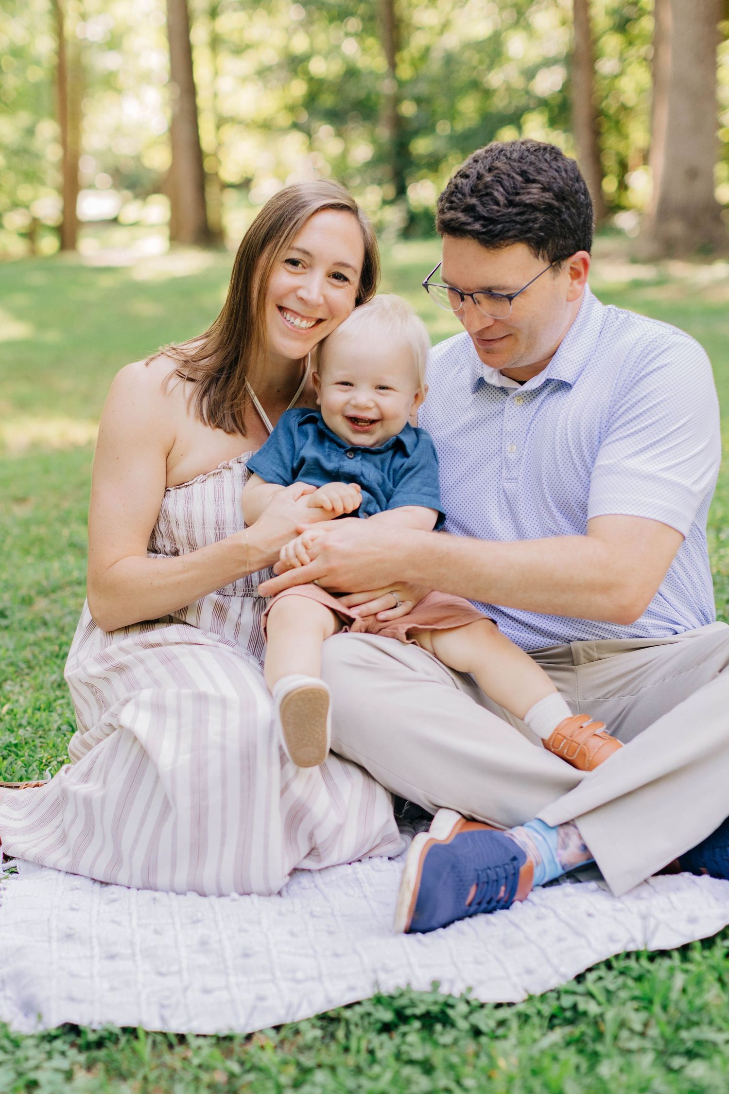 Happy young family with smiling baby boy sitting on blanket in green park during outdoor photo session.