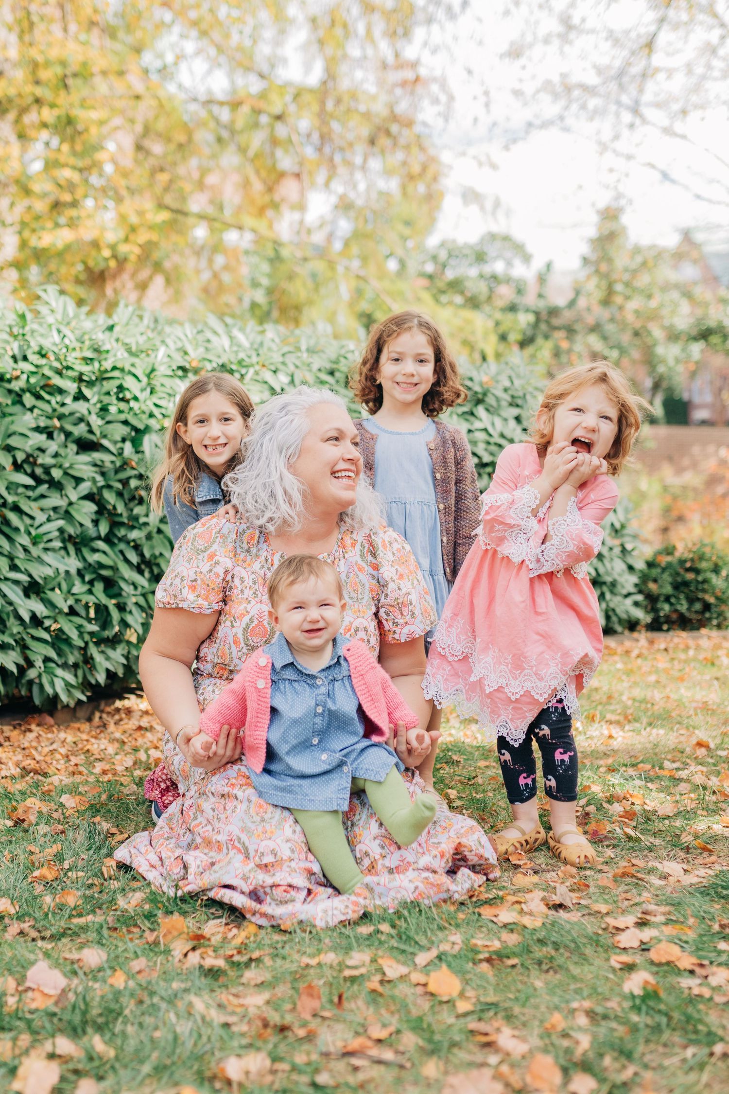 A grandmother sits on autumn grass laughing with four young grandchildren around her in a lush garden setting.