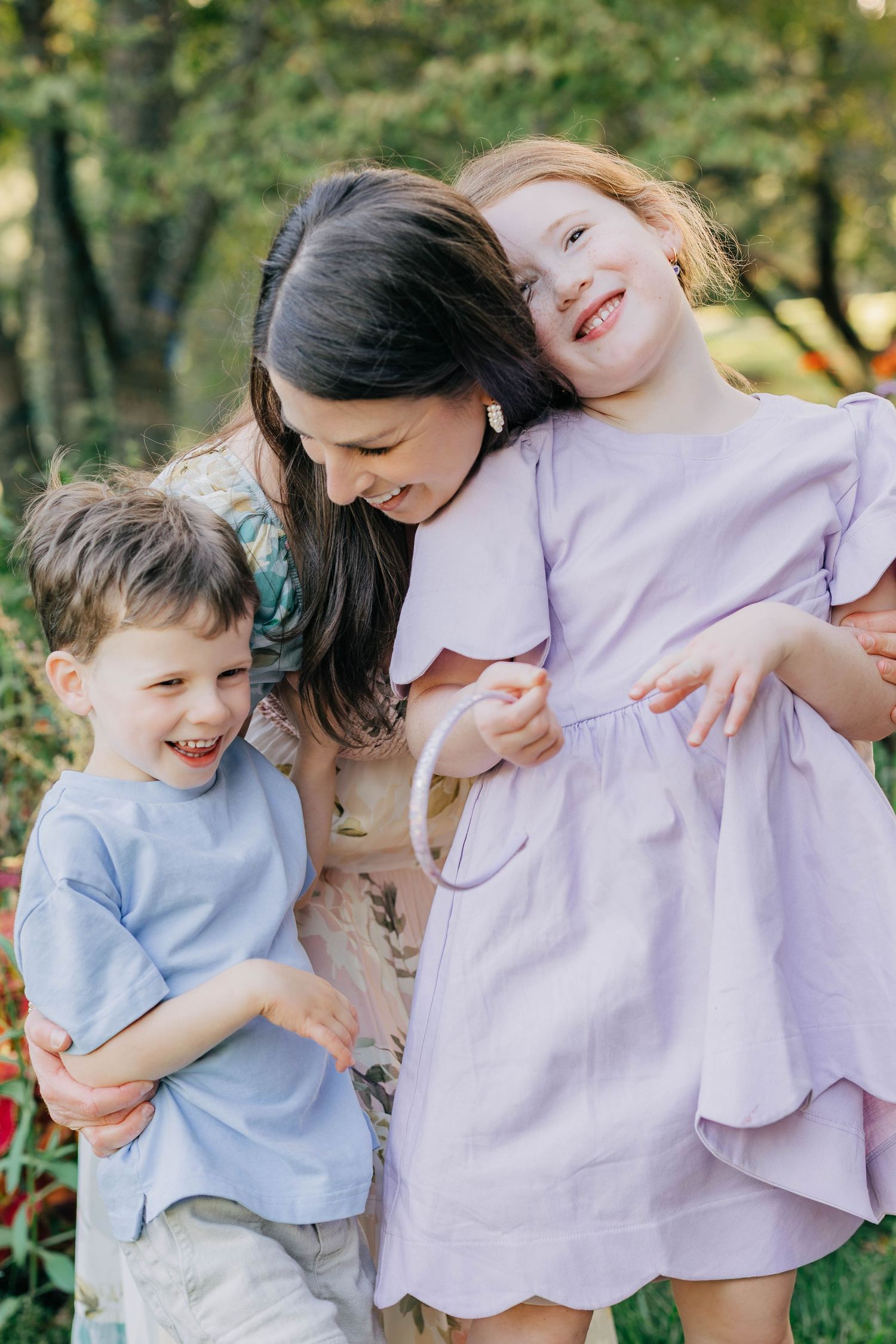 A mother laughs with her two young children outdoors in a lush green garden setting.