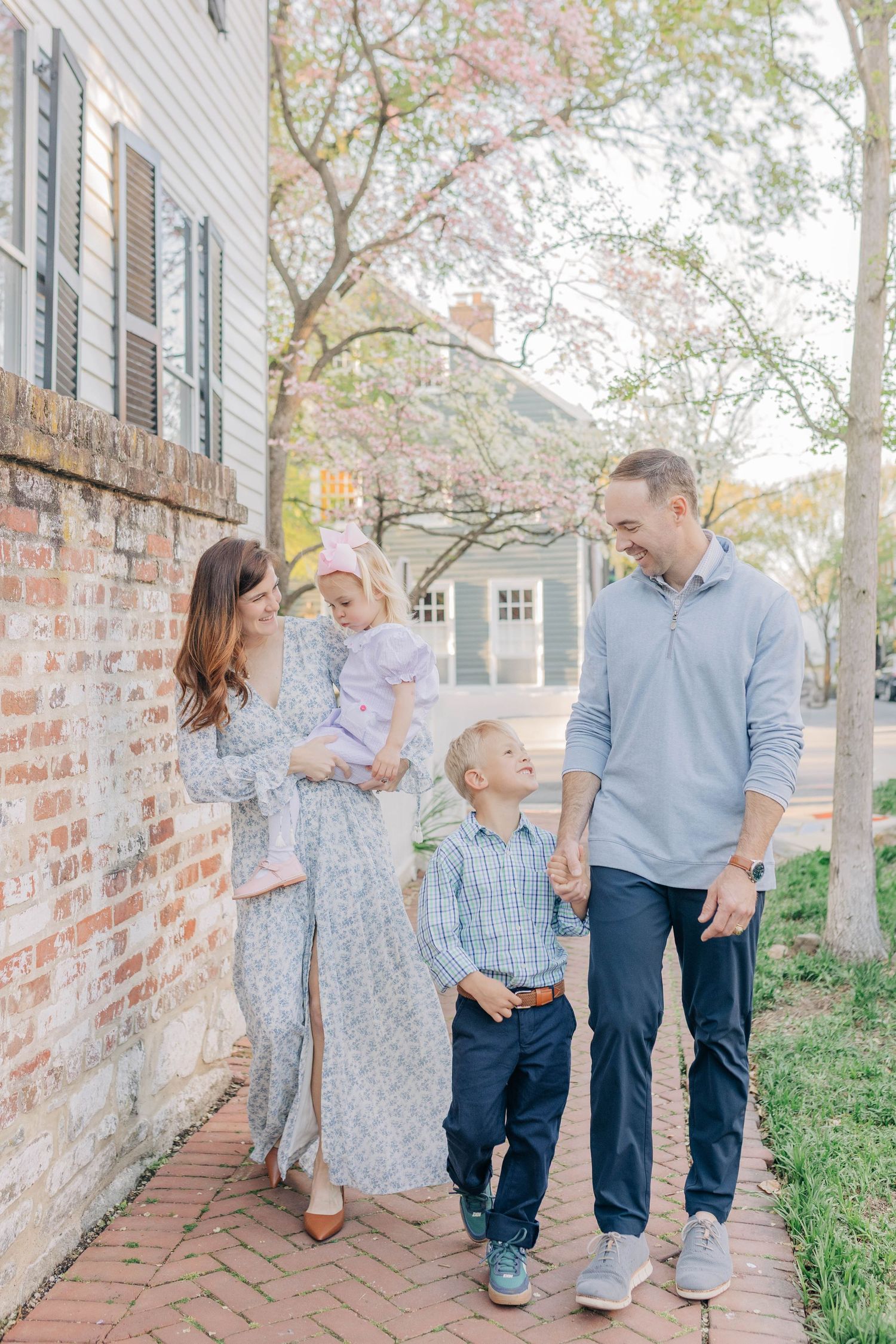 A happy family of four walks along a brick path beside a historic building with blooming trees in spring.