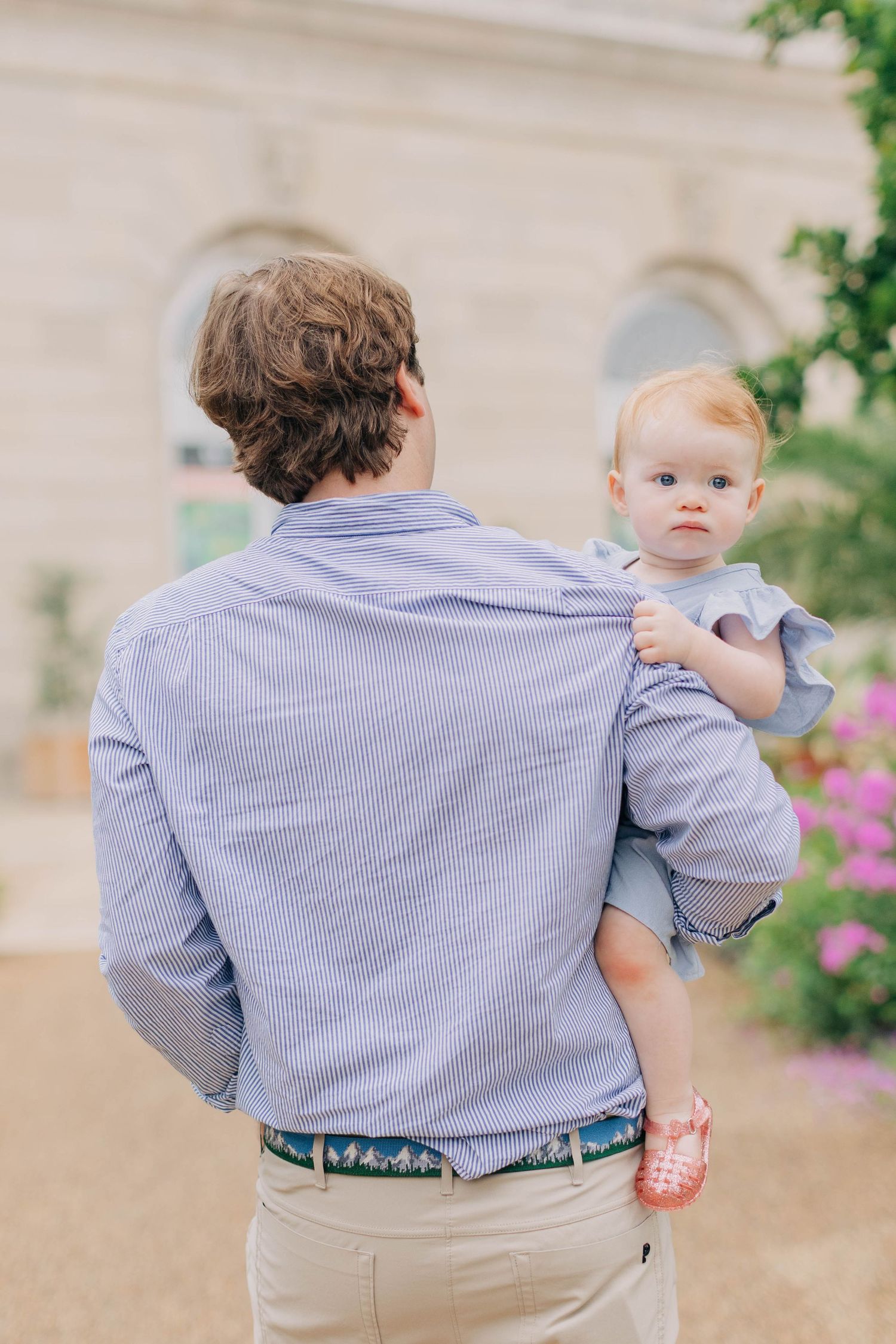 A father holds his baby girl in a blue dress outdoors near a garden.