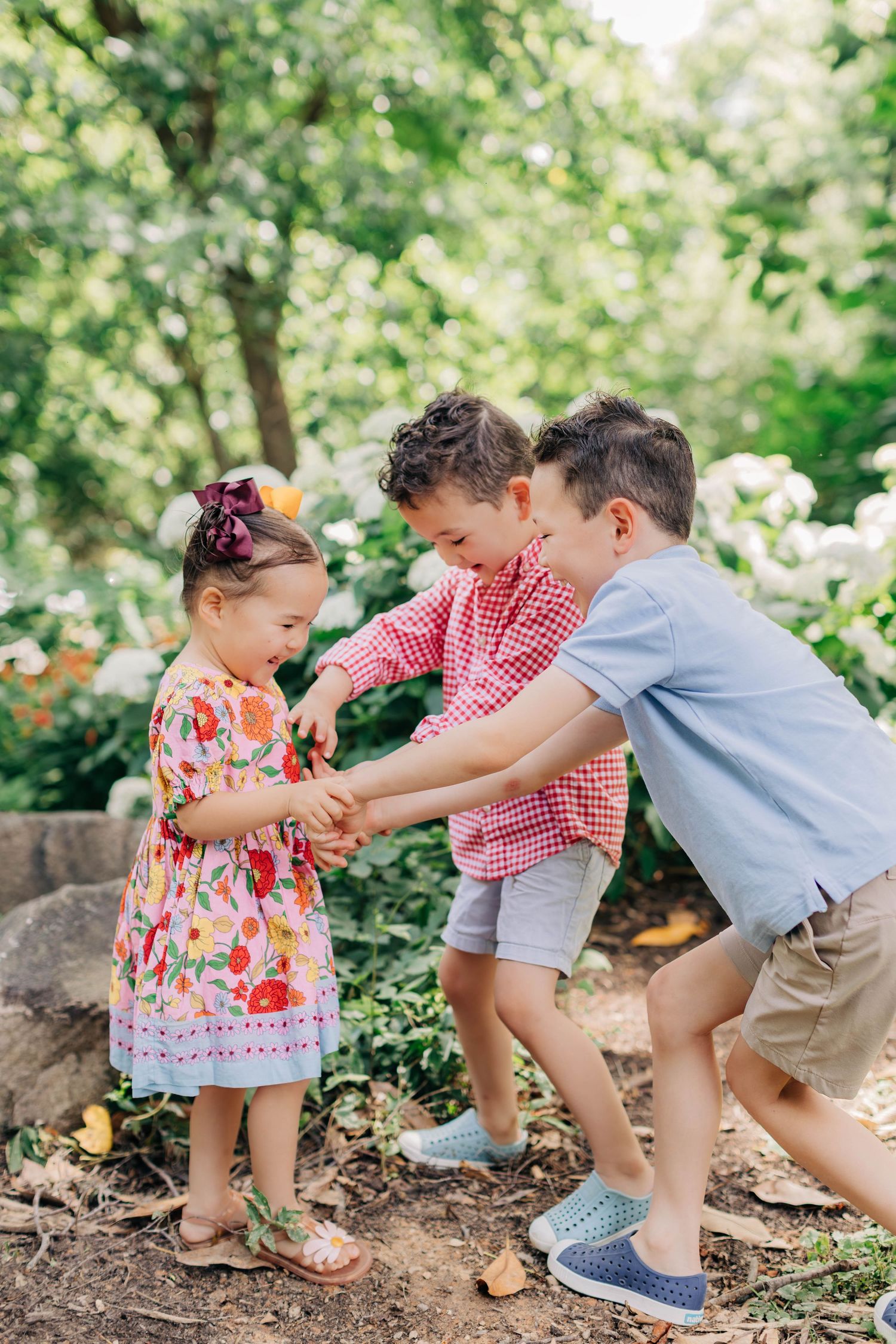 Three young children playing together in a lush garden, with a girl in a floral dress and two boys laughing outdoors.