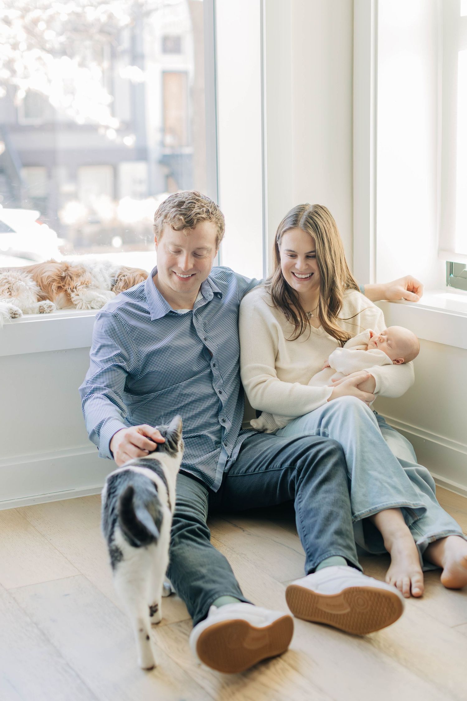 Happy parents sitting on floor with newborn baby and black and white cat by window.