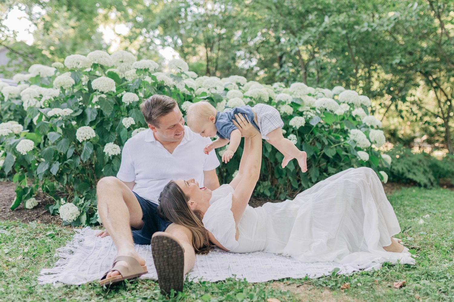 Family lying on white blanket in garden setting with flowering bushes.