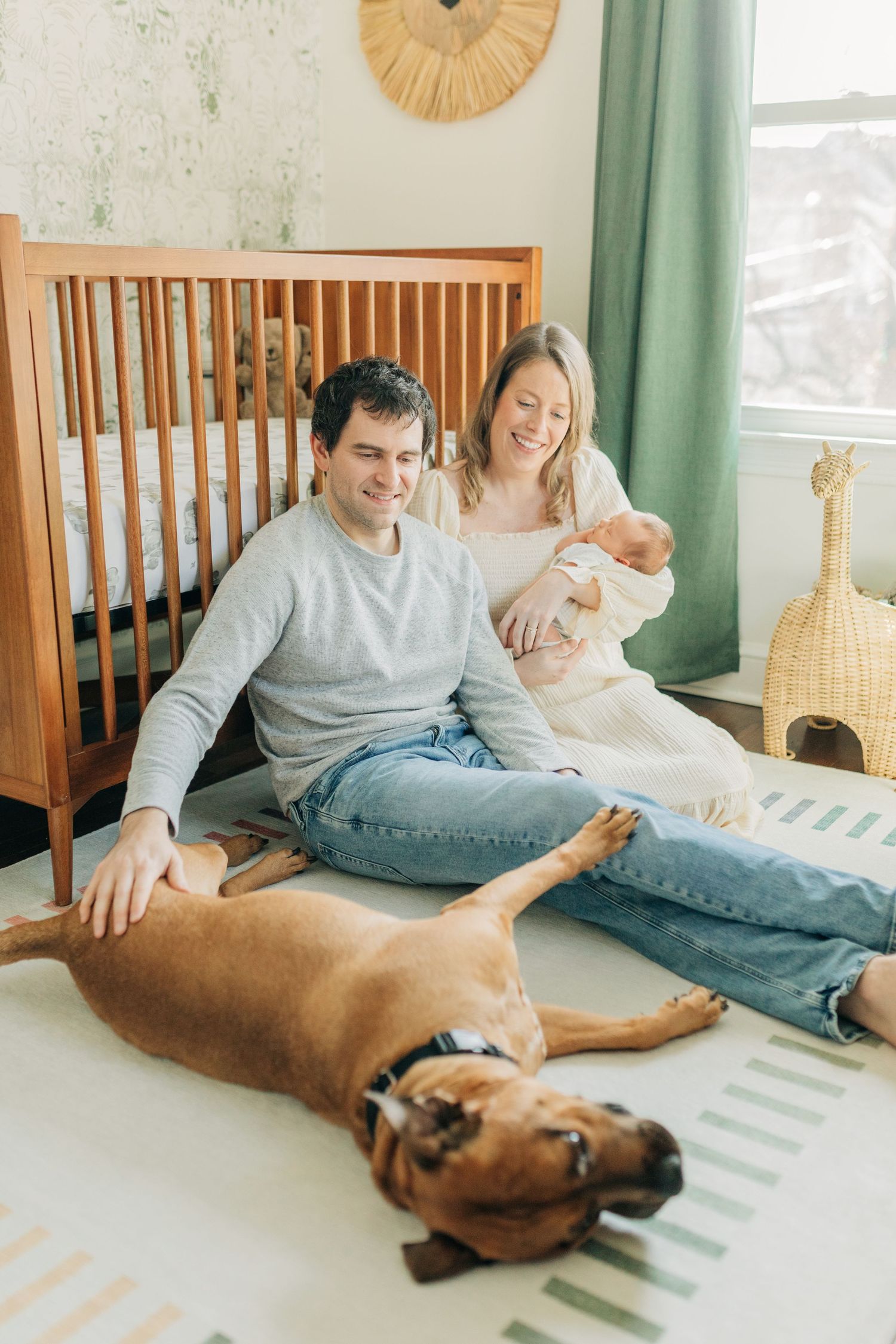 Family relaxes on floor with dog in nursery while holding newborn baby.