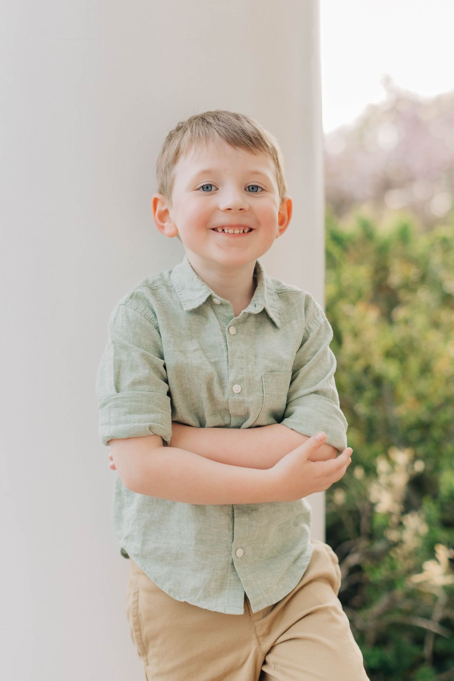 Smiling blond boy in green shirt and khaki shorts leaning against white pillar with arms crossed.