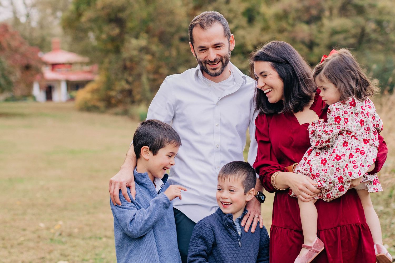 Happy family of five smiling outdoors in autumn park, parents with three young children in coordinated red and blue outfits.