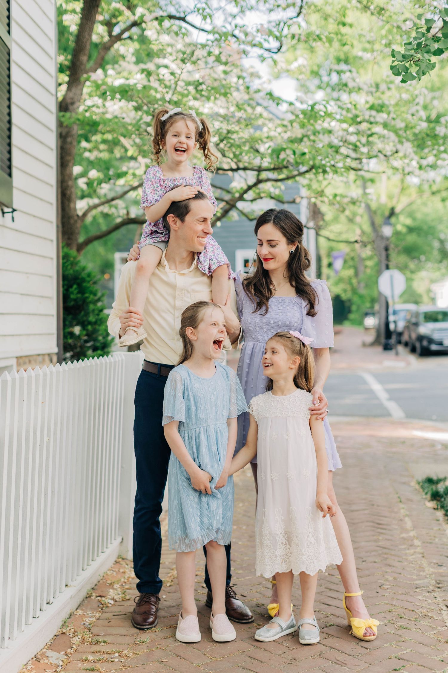 Happy family of five posing on a charming sidewalk, with two parents and three young girls in pastel spring dresses.