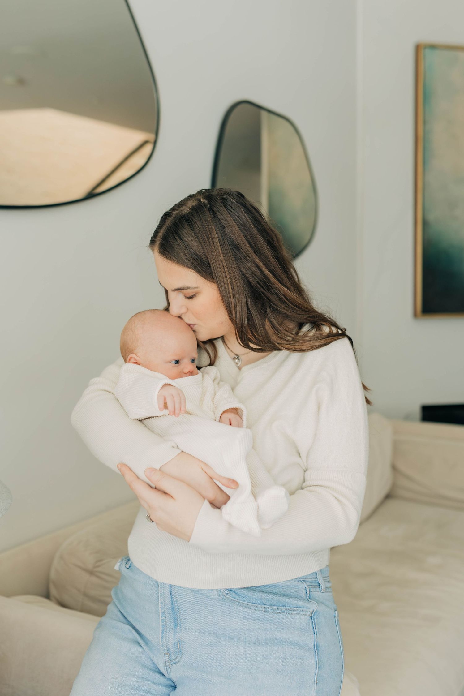 A young mother in a white sweater and jeans tenderly kisses her newborn baby dressed in white.