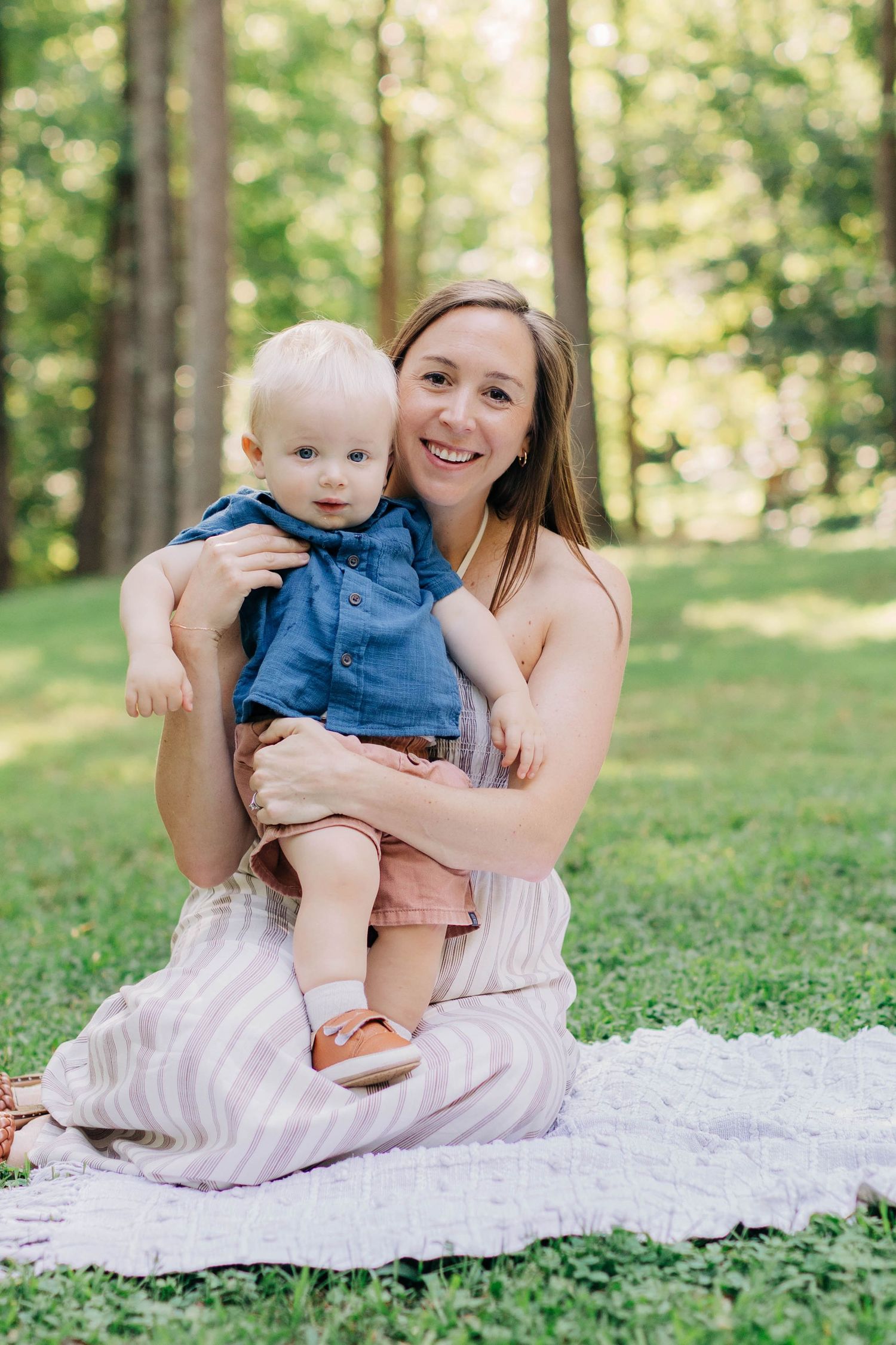 A smiling mother sits on a blanket in a green park, lovingly holding her blonde toddler in a denim shirt.