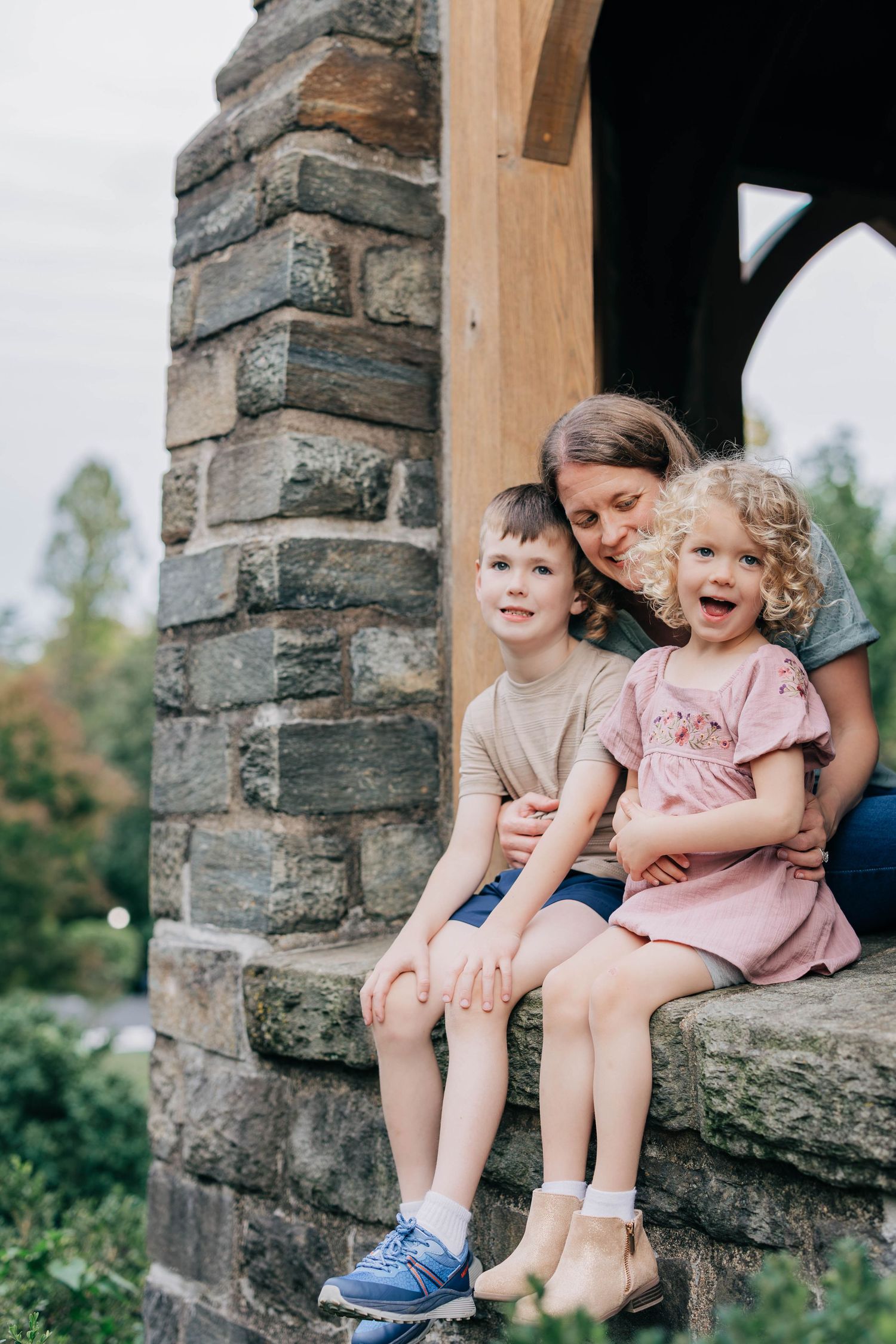 A smiling mother sits with her son and her laughing curly-haired daughter on a stone ledge beside a wooden post outdoors.