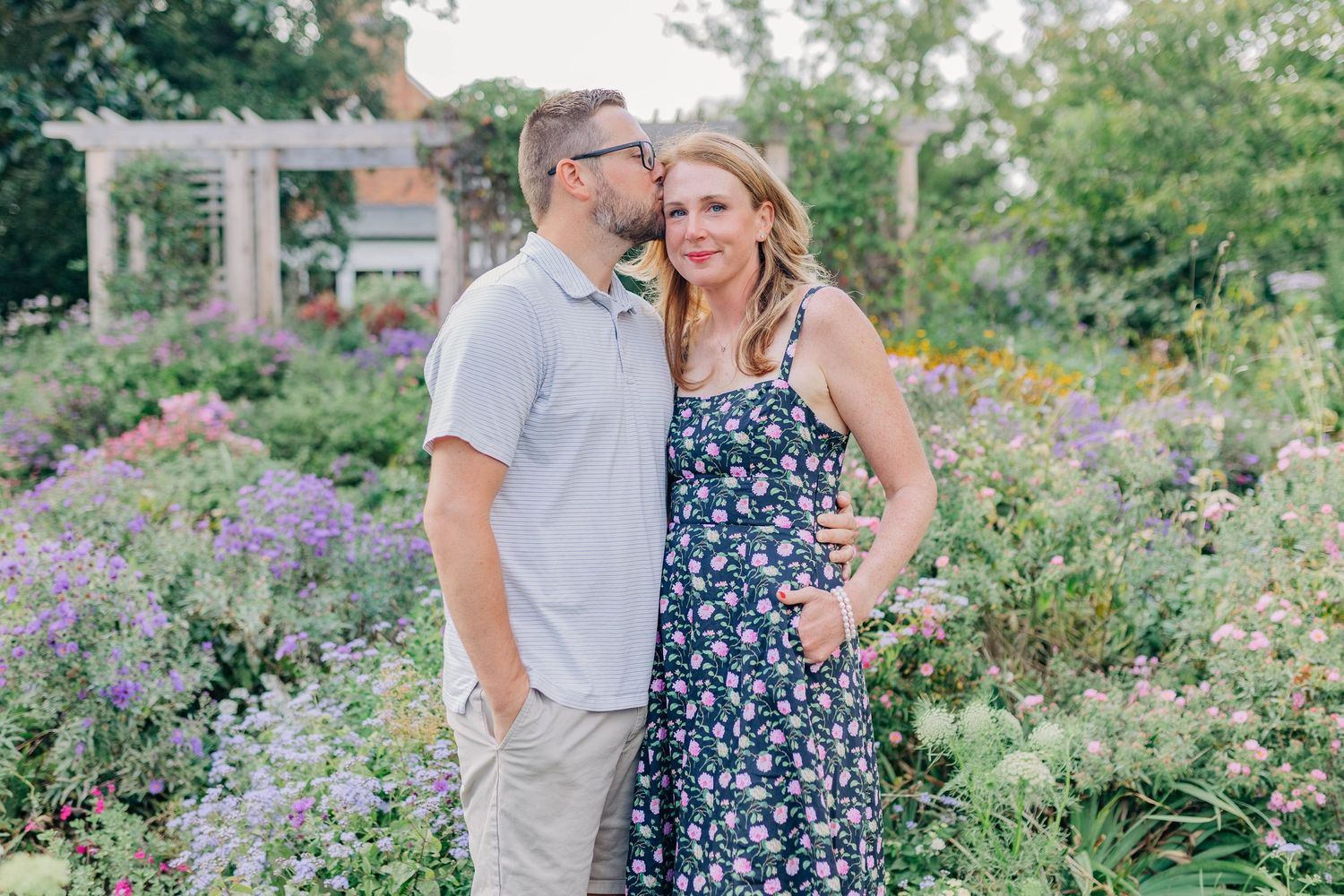 A couple poses together in a vibrant garden with purple flowers and a pergola in the background.