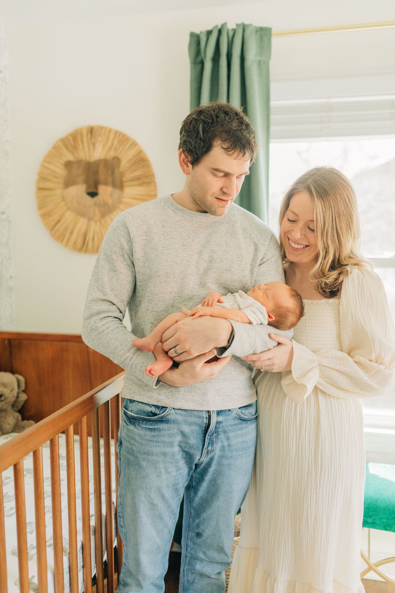 Parents stand by wooden crib holding newborn baby in nursery with green curtains and decorative wall art.
