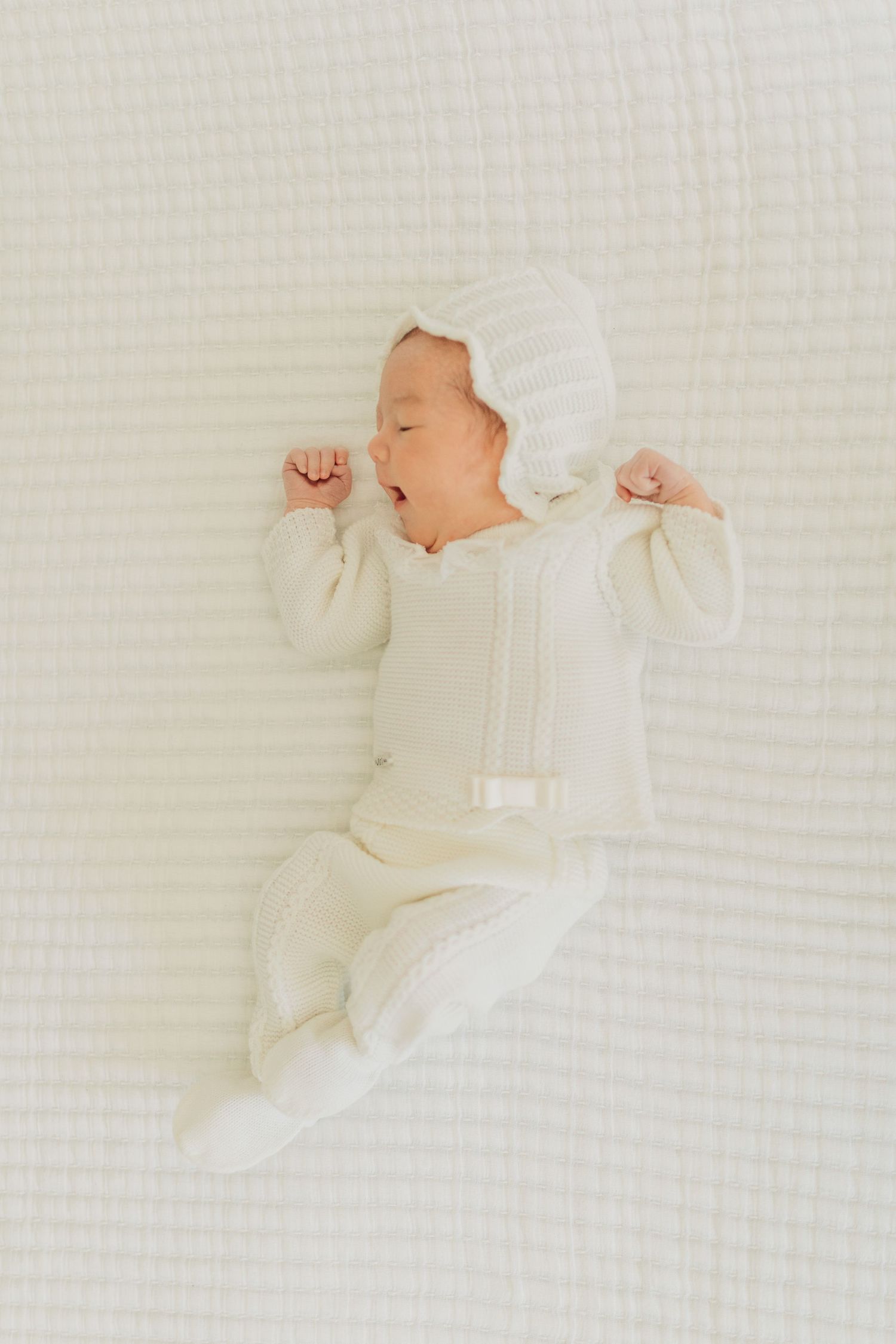 Baby in cream colored outfit sleeping peacefully on textured white bedding.