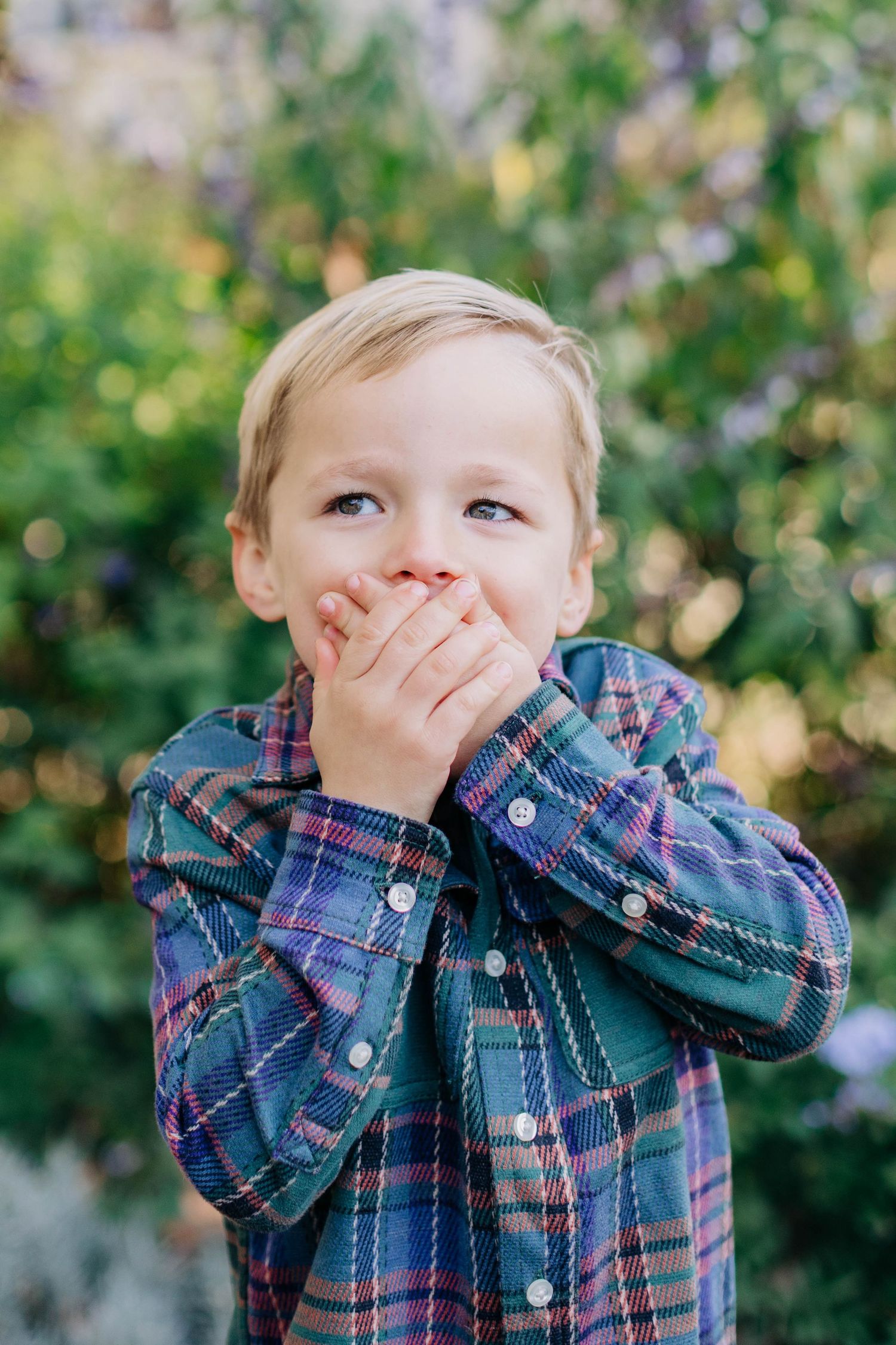 Child in plaid shirt making silly expressions while playing peek-a-boo outdoors.