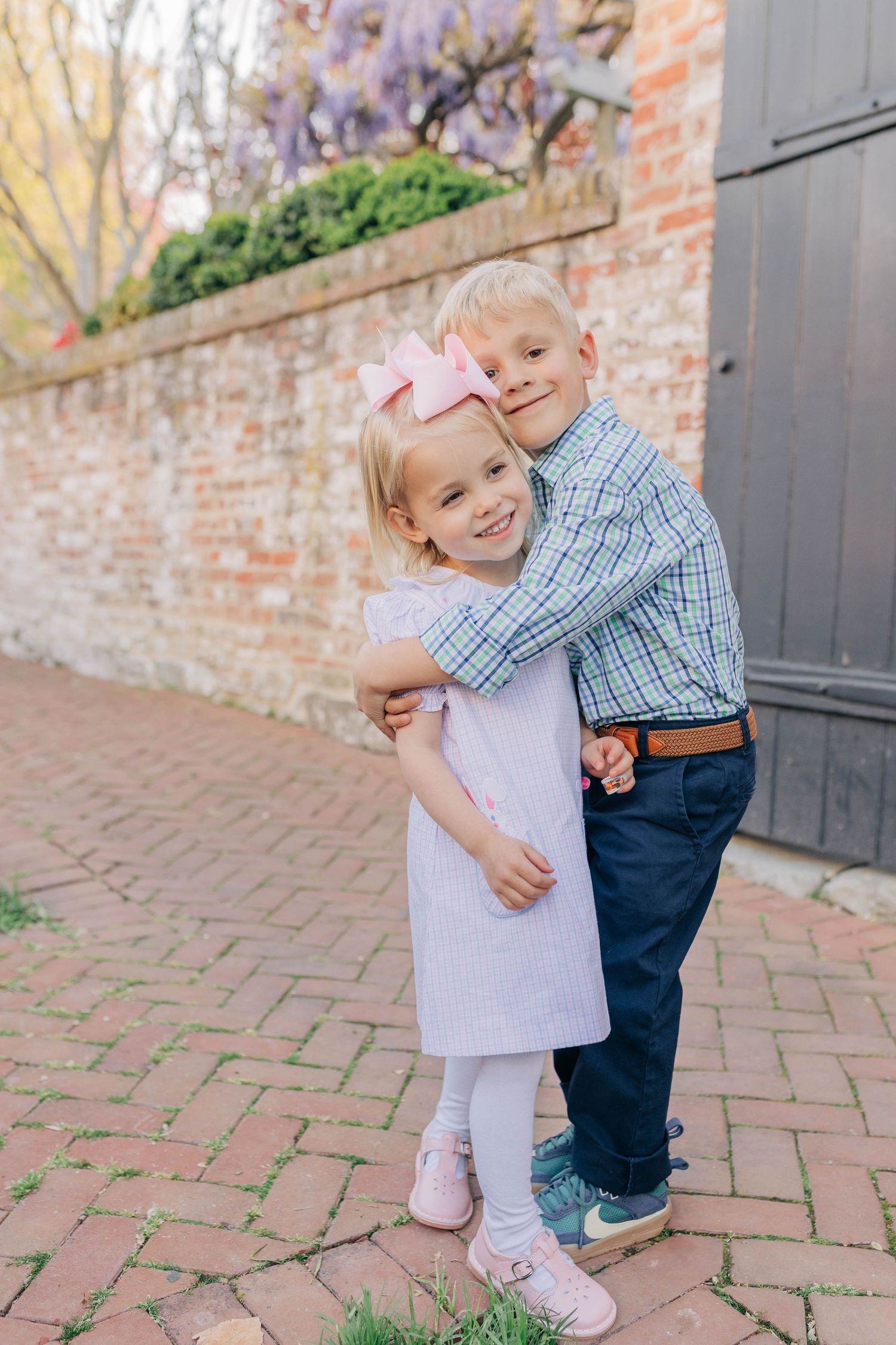 Two young siblings hug and smile outdoors near a brick wall, girl in pink dress with bow, boy in plaid shirt and jeans.